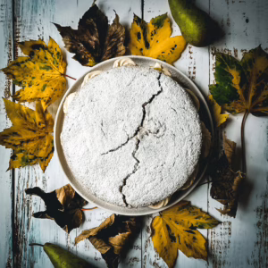 Chestnut and pear cake surrounded by fallen leaves and two pears, on a white wooden table