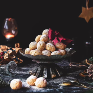 Soft amaretti piled up on a cake stand, surrounded by Christmas wrapping paper, dried orange slices, pinecones and cardboard stars hanging from the ceiling