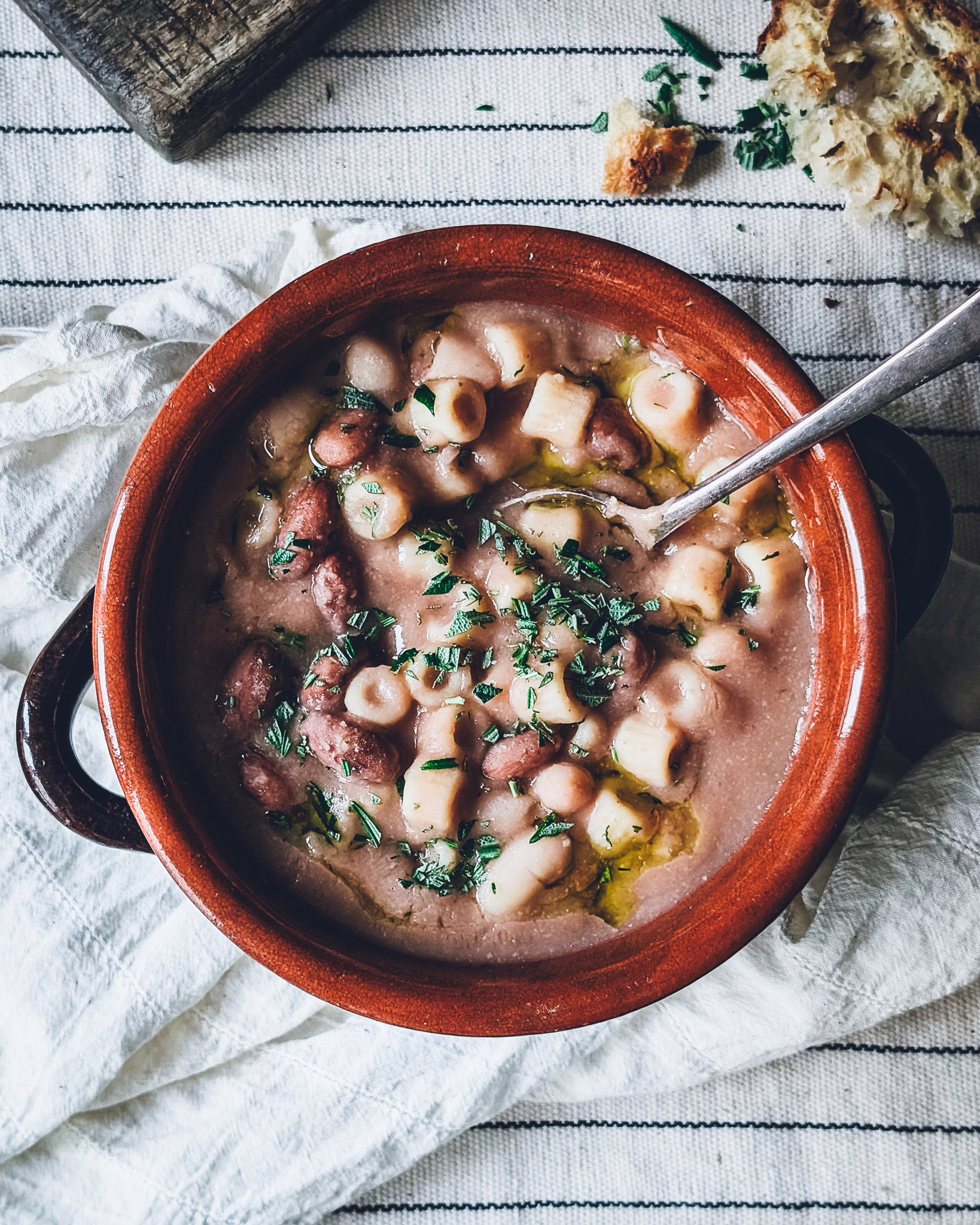 Rustic terracotta bowl full of pasta e fagioli with some bowls and crusty bread in the background
