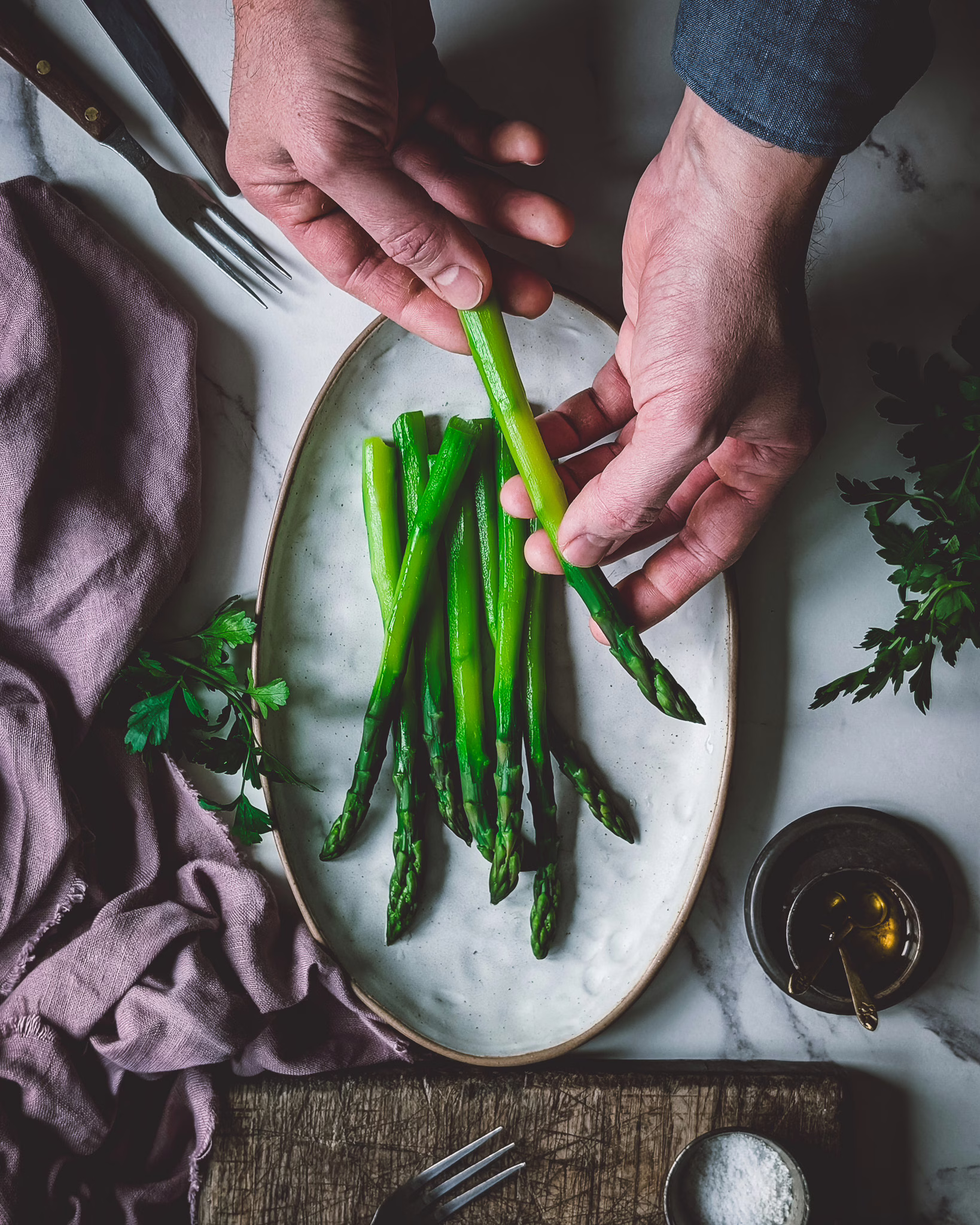 Hands of person placing one cooked green asparagus on a plate with more asparagus. The plate is beside a vintage purple cloth and a wooden chopping board, on a white marble background.