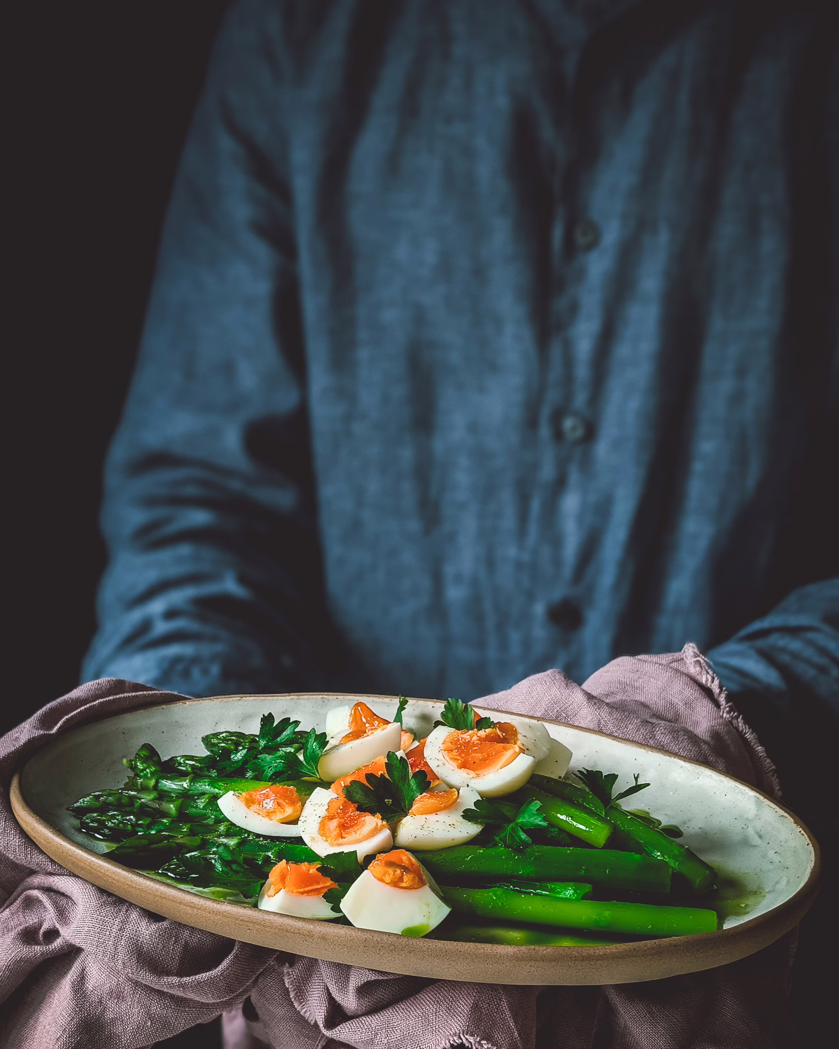 Person wearing blue shirt and carrying a vintage purple cloth with a ceramic plate with asparagus and boiled eggs on top.