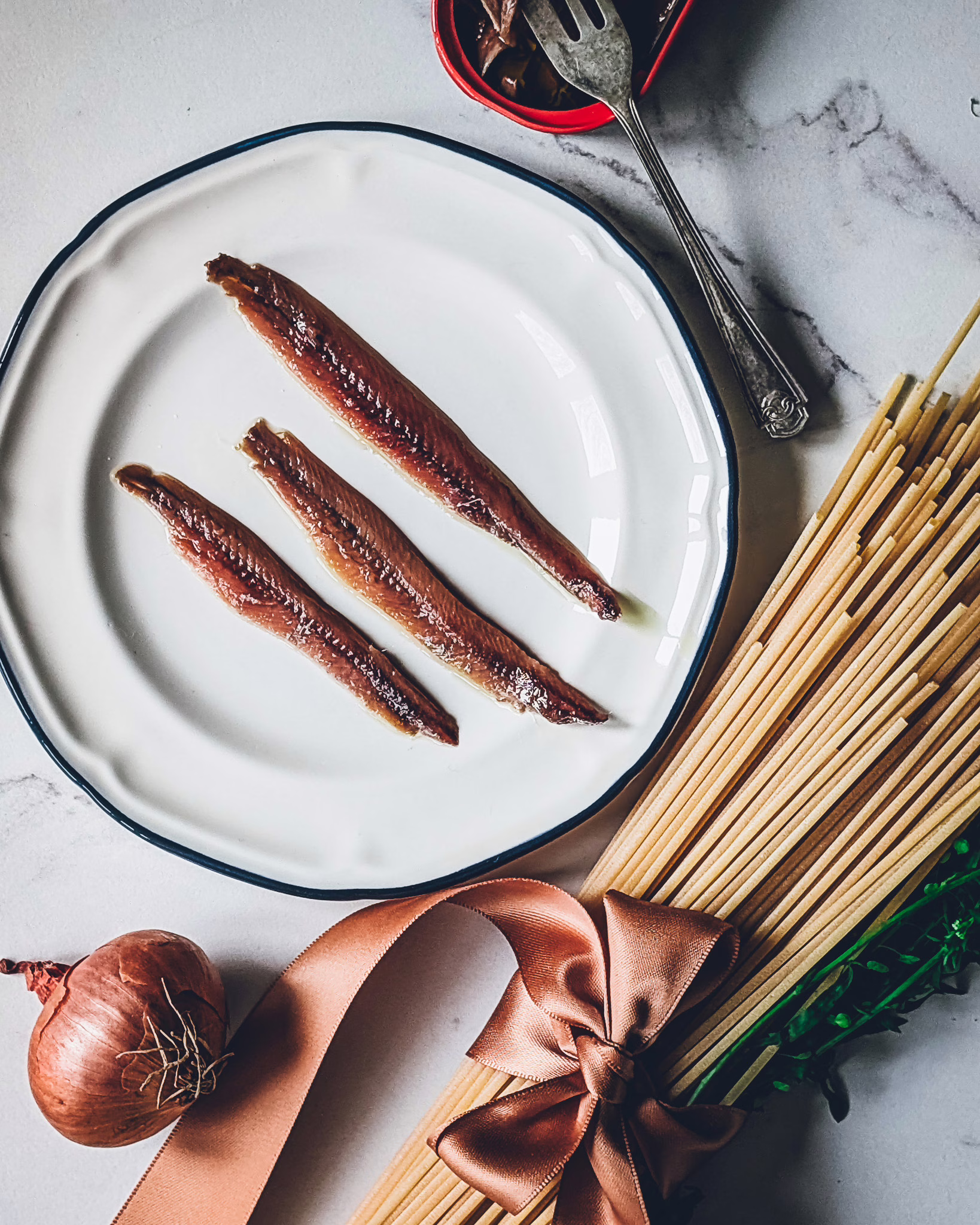 Close up of a small plate with three anchovies on a marble table. On the table there is also an open can of anchovies, a small fork , a dry wooden branch and some spaghetti and wild herbs kept together with a brown ribbon