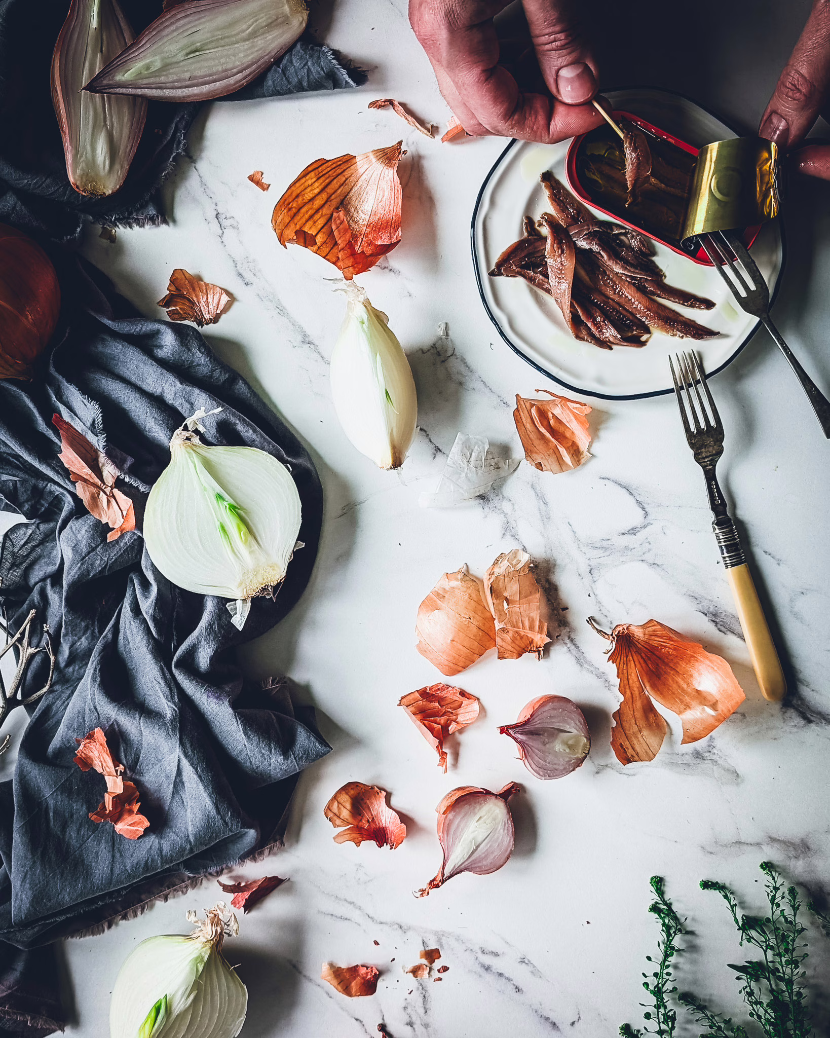 Sliced onions on a marble table alongside a blue cloth and small plate with a can of anchovies on it, and some anchovies out of the can. Cutlery and small trinkets are also on the table, and a man is taking an anchovy out of the can.