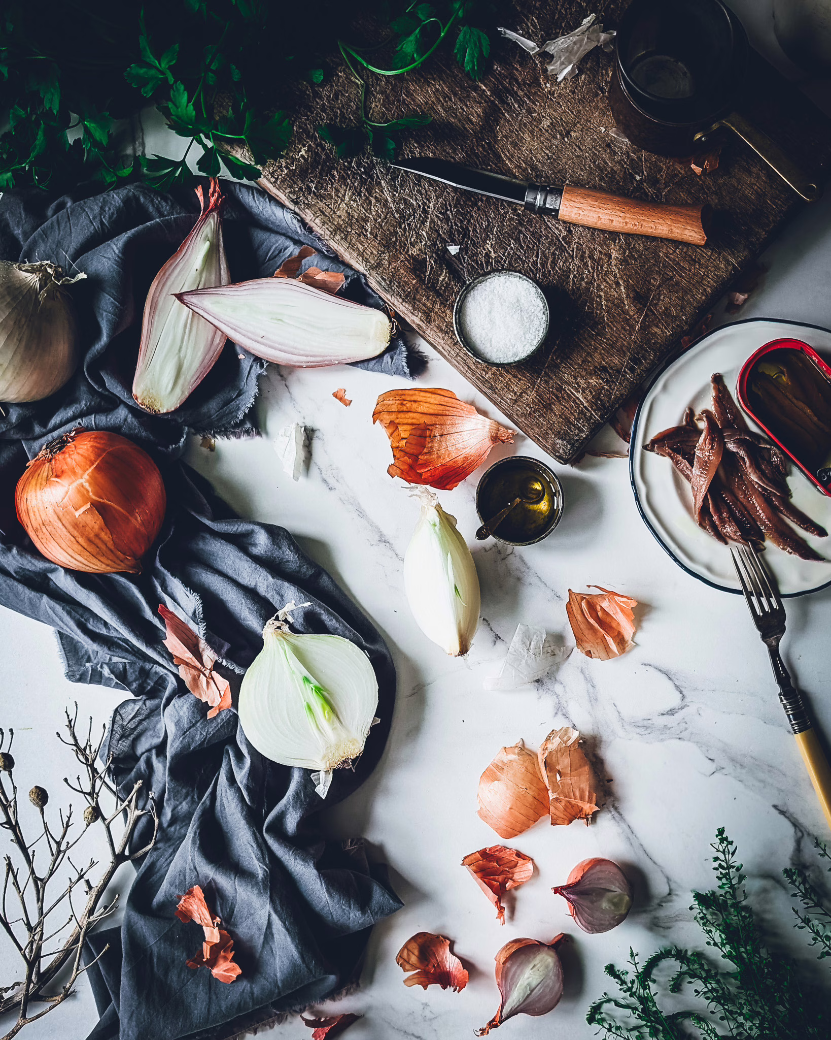 Sliced onions on a marble table alongside a wooden chopping board, a blue cloth and small plate with a can of anchovies on it, and some anchovies out of the can. Cutlery and small pans are also on the table, as well as a bunch of parsley.