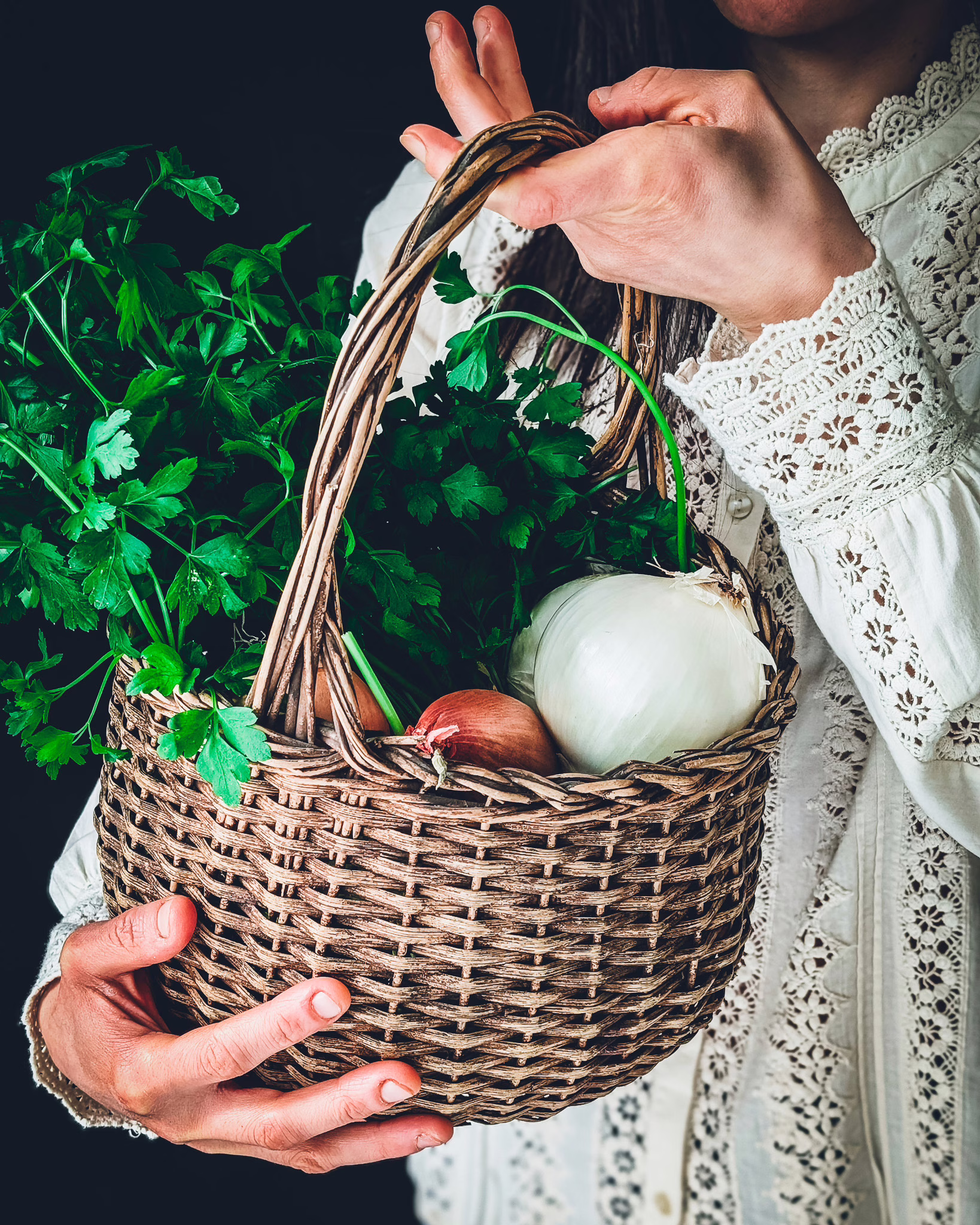 Woman wearing a vintage embroidered white shirt holding a basket full of parsley and onions of different variety and colour
