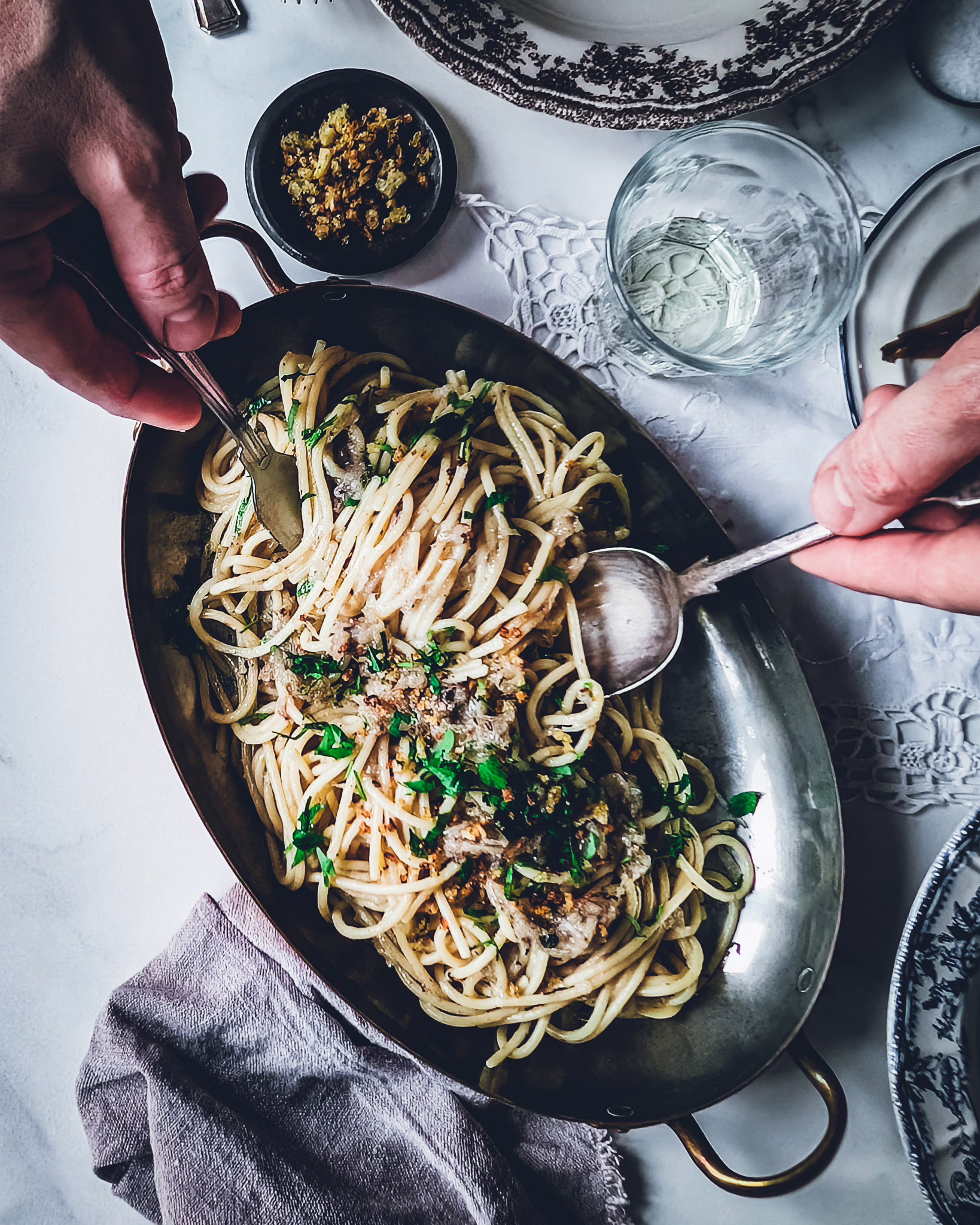 Hands of a person serving pasta using fork and knife from a serving plate full of bigoli in salsa