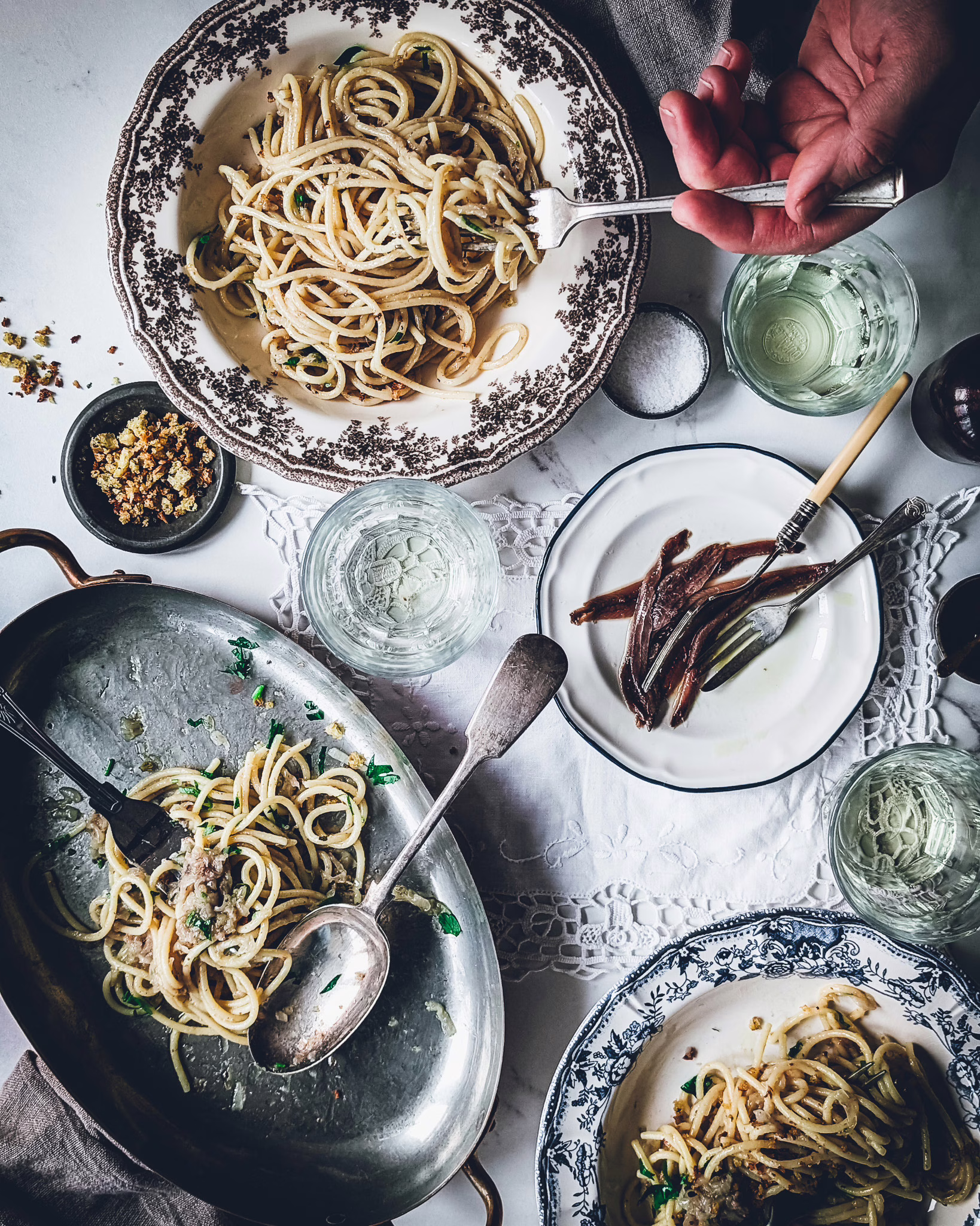 Serving plate of pasta half empty, next to two plates full of pasta, on a table with a small plate of anchovies, glasses with white wine and trinkets. A hand is seeing using a fork to eat pasta from one of the dishes.