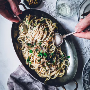 Hands of a person serving pasta using fork and knife from a serving plate full of bigoli in salsa