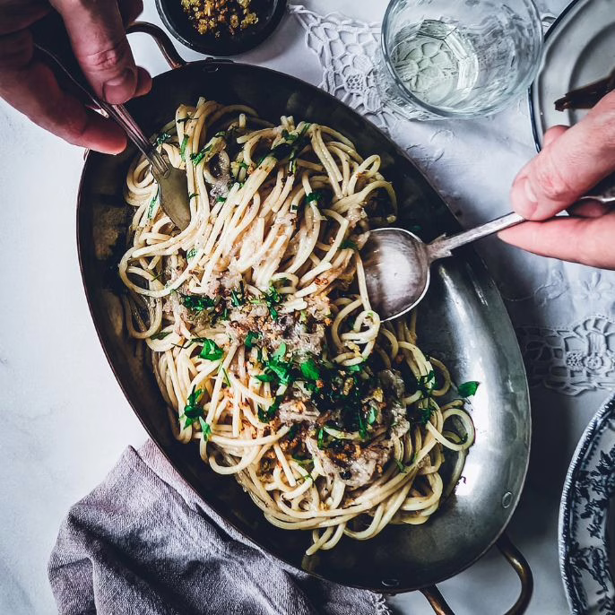 Hands of a person serving pasta using fork and knife from a serving plate full of bigoli in salsa