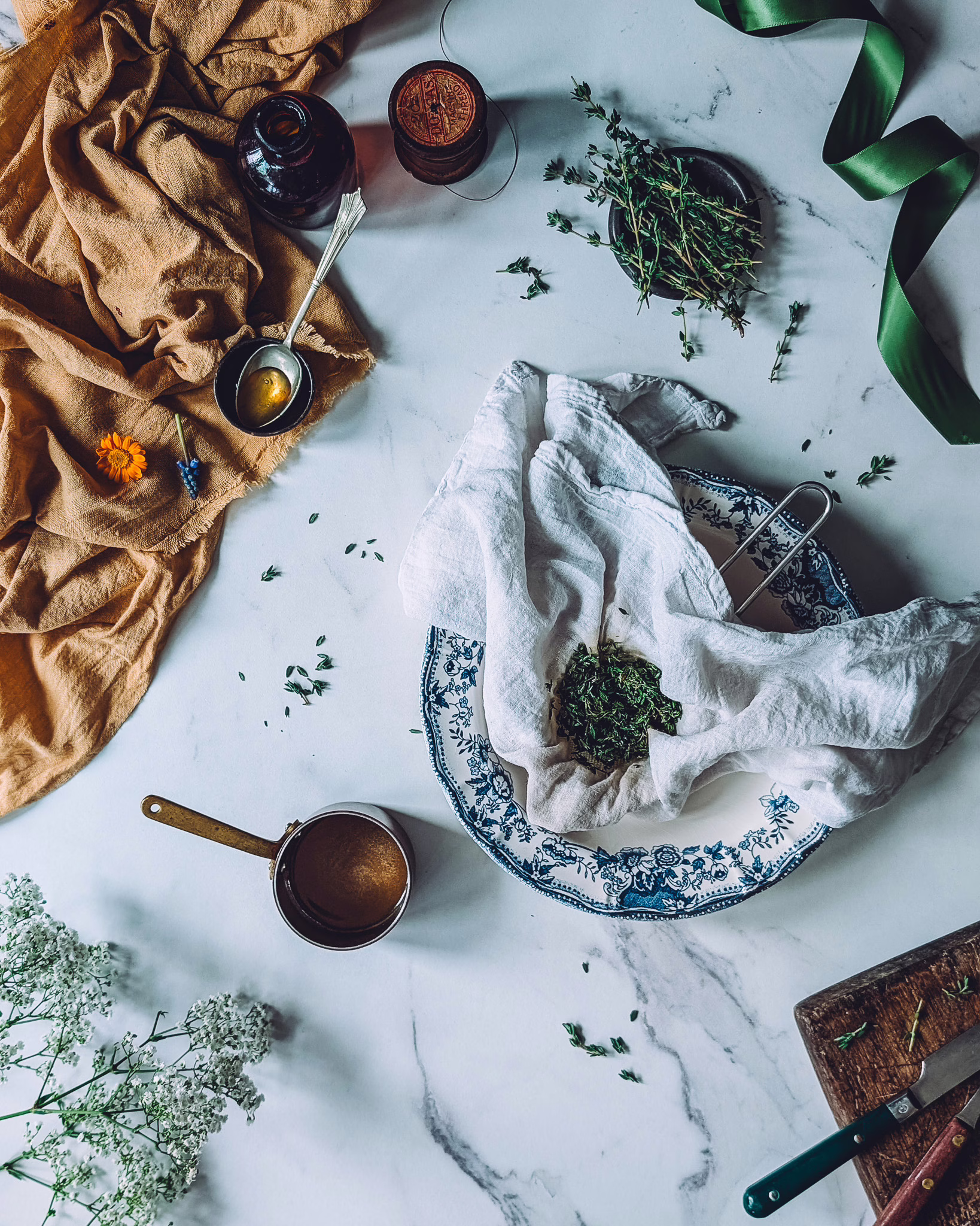 Thyme honey being filtered through muslin cloth on plate with blue vintage rim. The plate is on a marble table, next to a small pot with melted honey, a green ribbon, some line cloth, fresh thyme, vintage cutlery and a brown glass bottle.