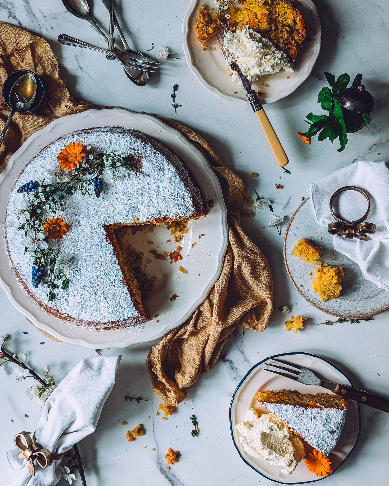 Serving plate with Italian carrot cake missing two slices. The slices are on two small plates alongside some whipped cream. The marble table is decorated with flowers, cutlery and linen towels.