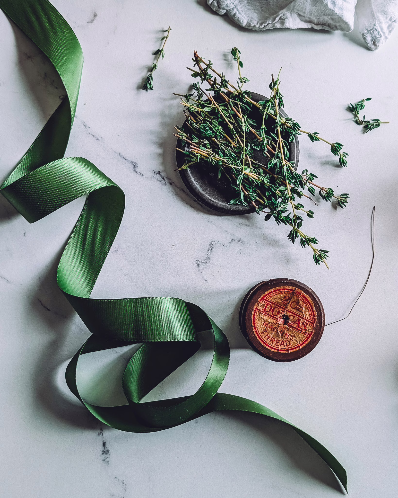 Green ribbon, a spool of thread and a small vessel with fresh thyme on a marble table