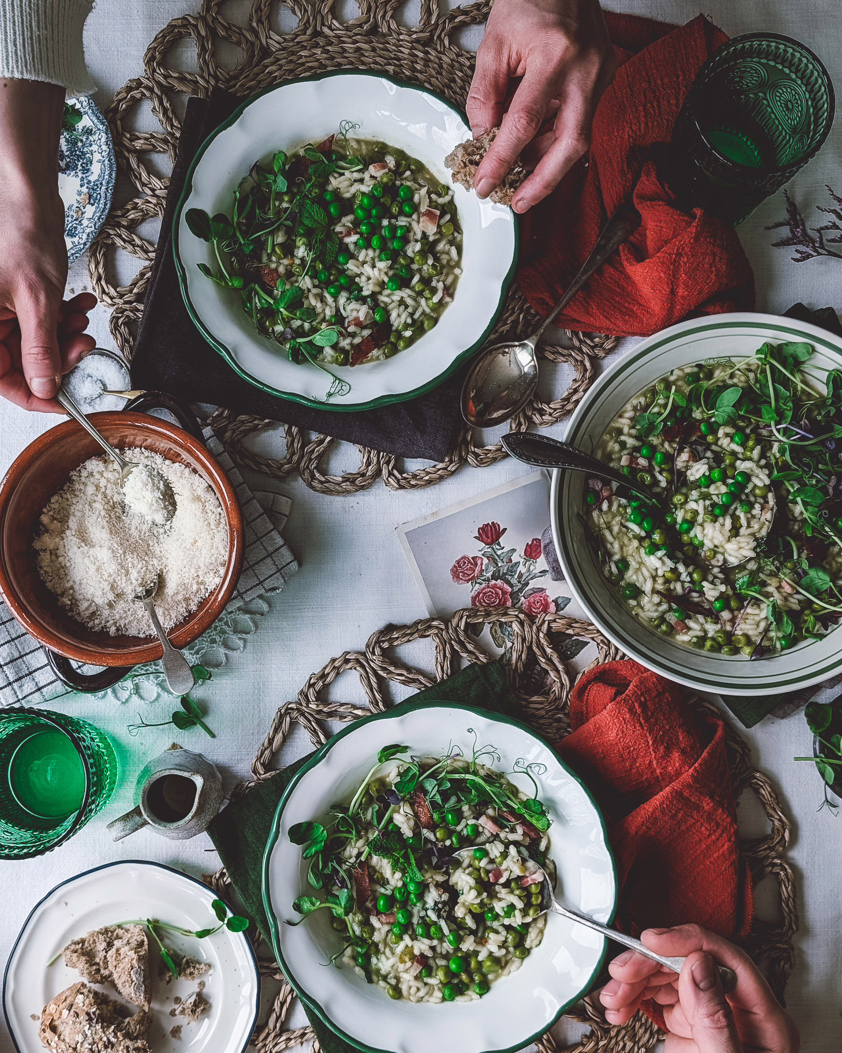 Two people eating rice and peas. The plates are on a table decorated with terracotta napkins, rattan chargers, green glasses, a serving bowl, and a terracotta bowl full of grated parmesan.