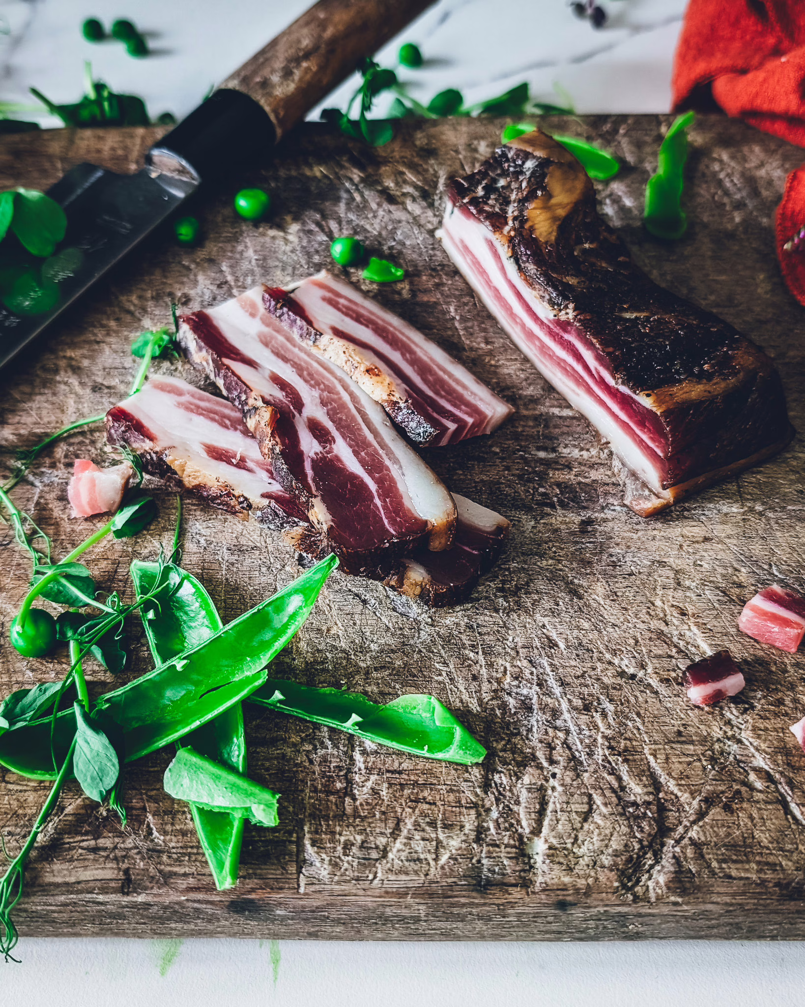 Slices of pancetta on a wooden chopping board. Pea shoots, pea pods and a knife are also visible in the picture.