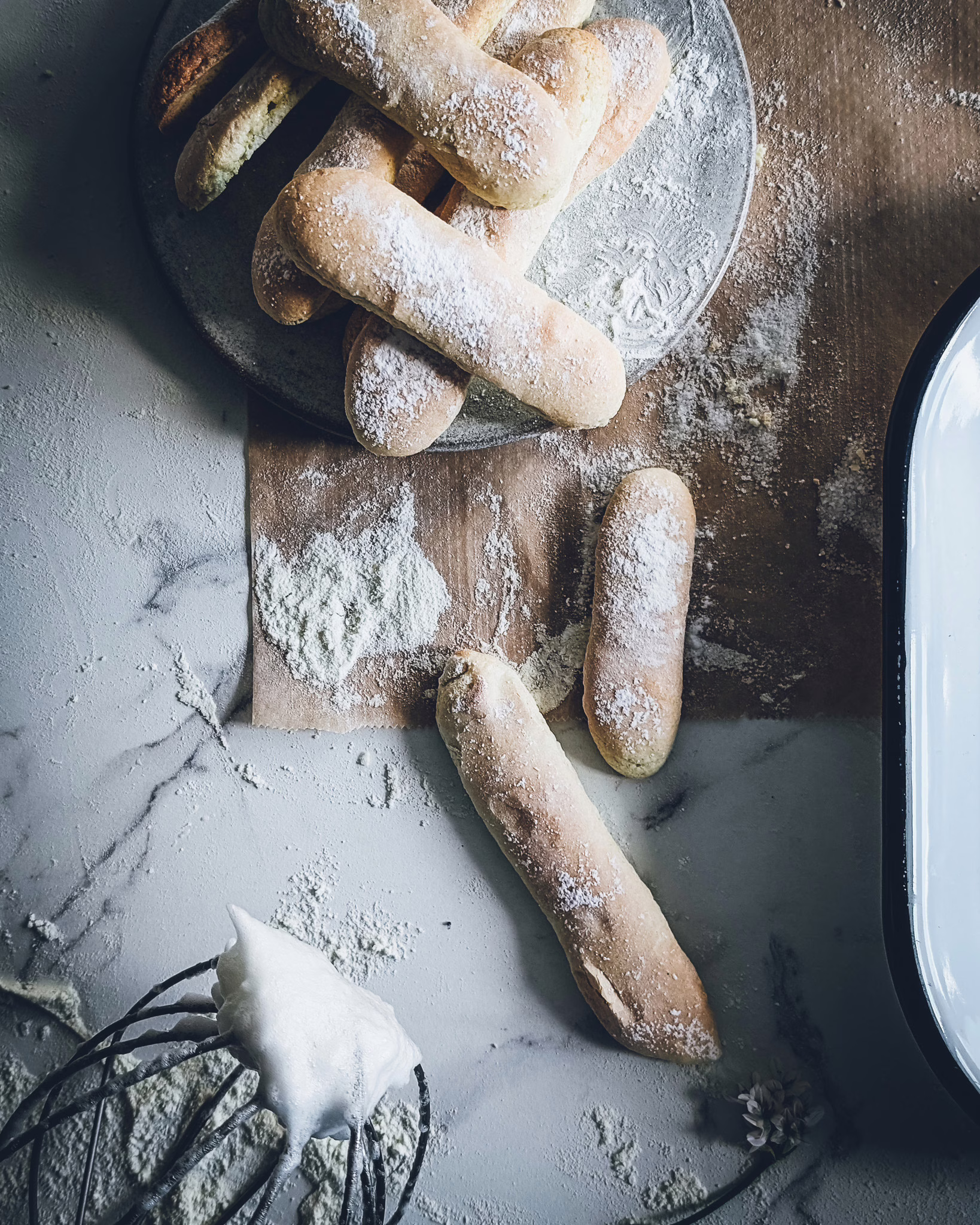 Ladyfingers on a plate and on the marble table. On the table also a whisk attachment with some egg white on it.