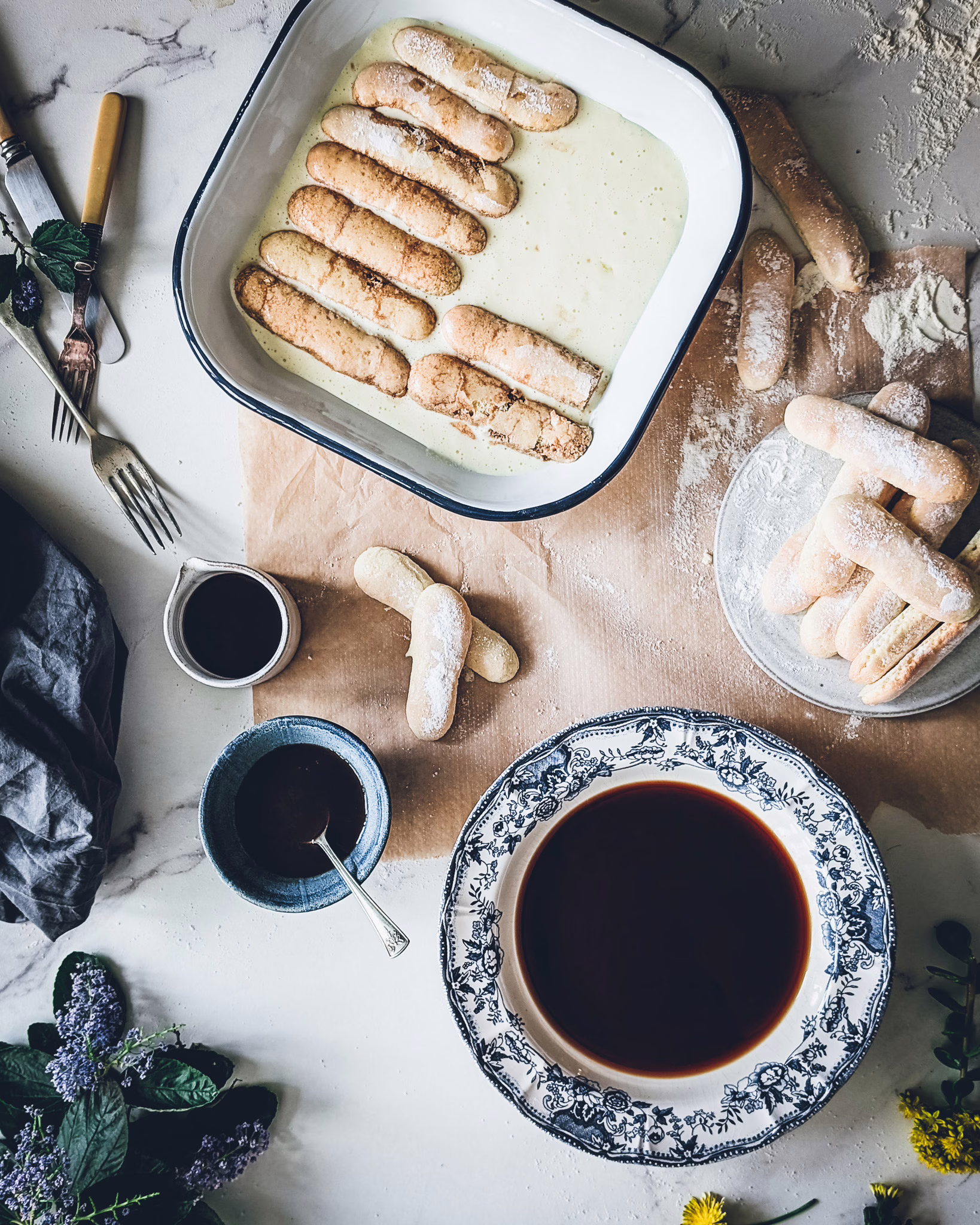Tin with mascaprone cream and dipped ladyfingers. On a small plate some ladyfingers. Also on the table some mugs and a plate with coffee.