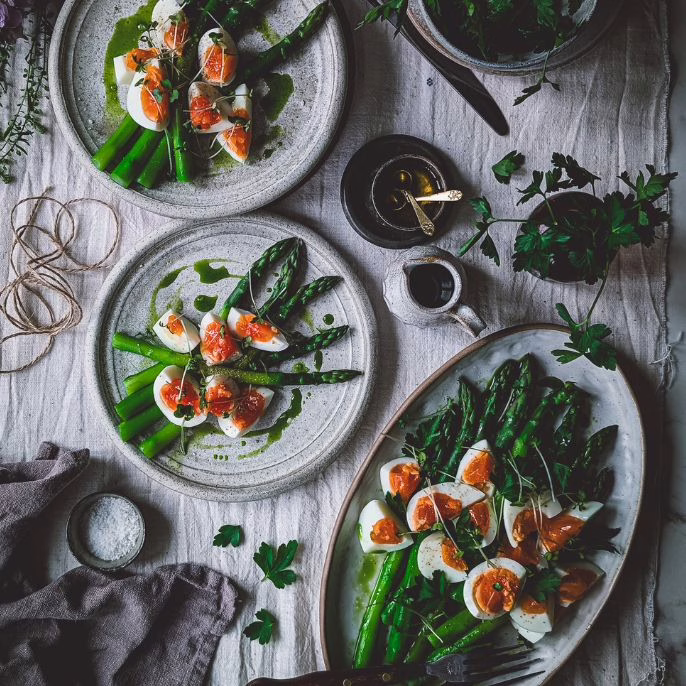 Serving plate with asparagus and boiled eggs. Two plates full of asparagus and eggs are also beside the plate on the table, alongside rustic cutlery and trinkets with oil, plus a small bottle with parsley leaves.