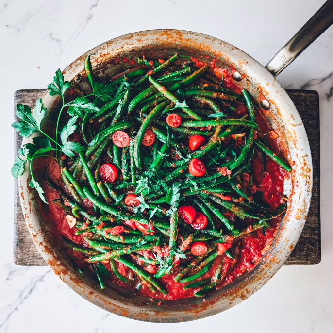 Pan with green beans in garlicky tomato sauce on a wooden chopping board