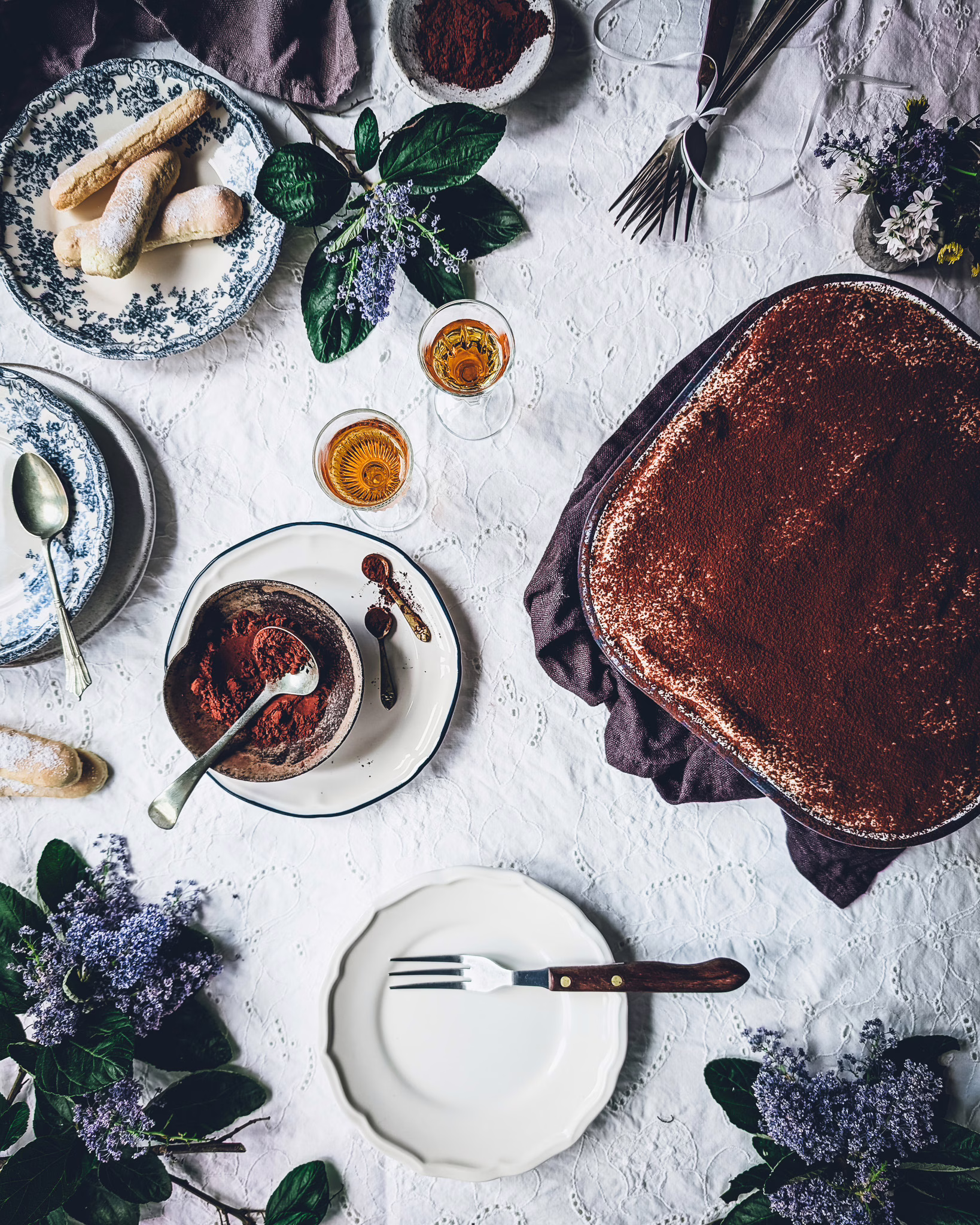 Empty small plate. On the table a tin with some eaten tiramisu, some plates with cocoa powder, a plate with ladyfingers, another plate with a tiramisu slice, some vintage cutlery and some purple flowers.