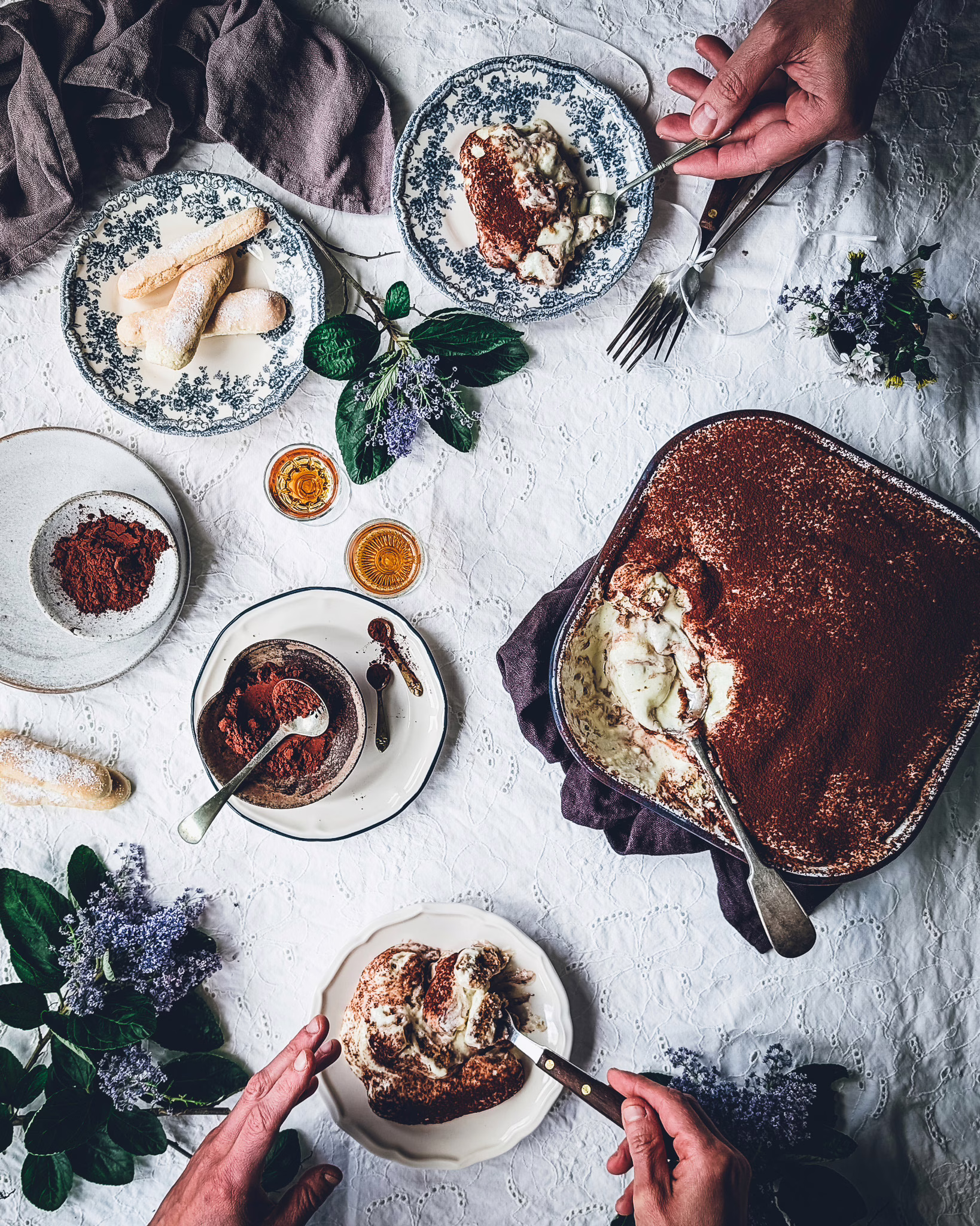 Person eating tiramisu from a small plate. On the table a tin with some eaten tiramisu, some plates with cocoa powder, a plate with ladyfingers, another plate with a tiramisu slice, some vintage cutlery and some purple flowers.