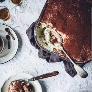 Tin of tiramisu partially eaten. A large serving spoon is in the tin. On the side a plate with tiramisu on it.