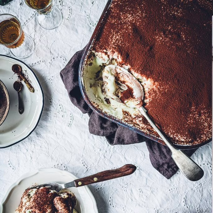 Tin of tiramisu partially eaten. A large serving spoon is in the tin. On the side a plate with tiramisu on it.