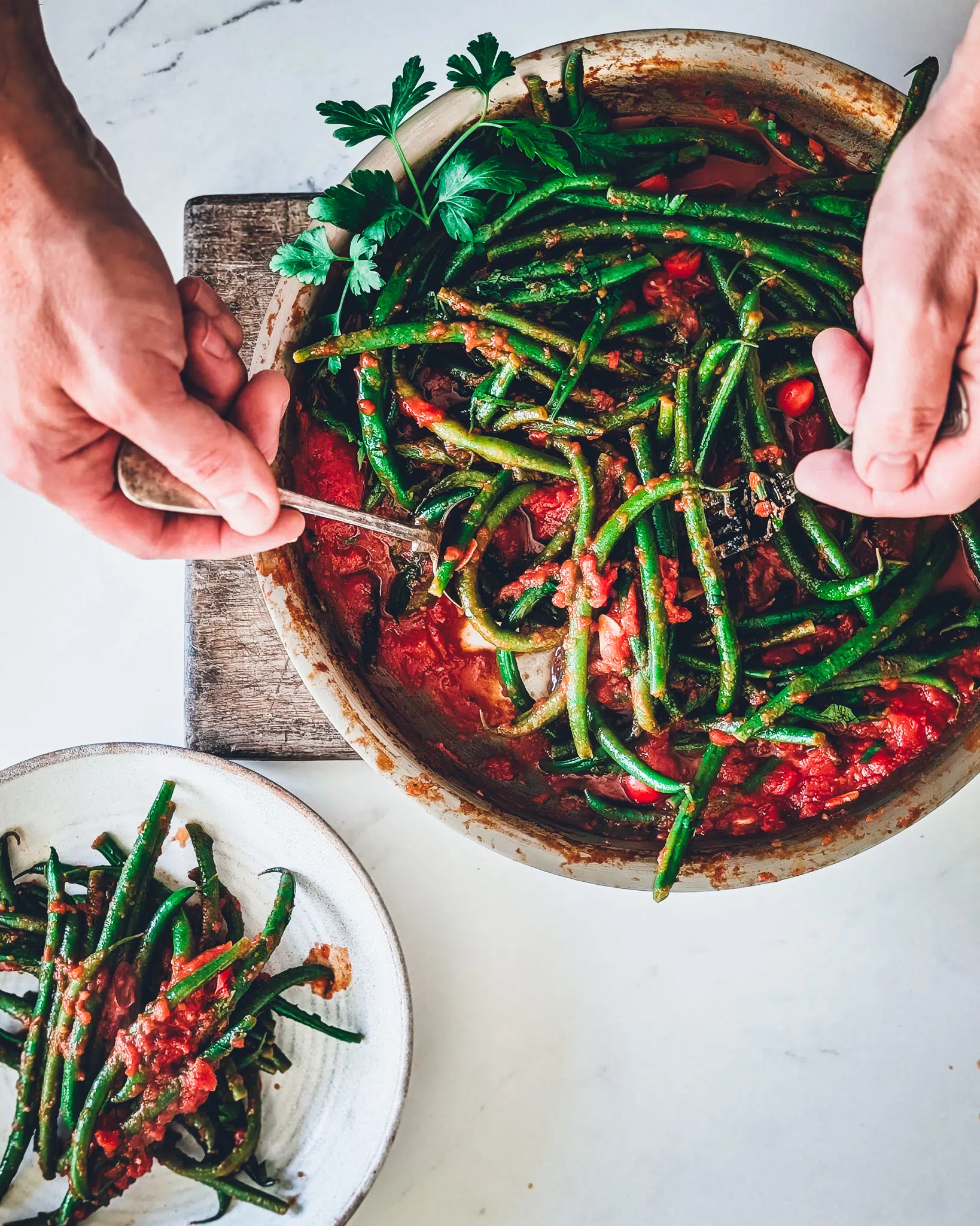 Person serving from a pan with green beans in garlicky tomato sauce on a wooden chopping board