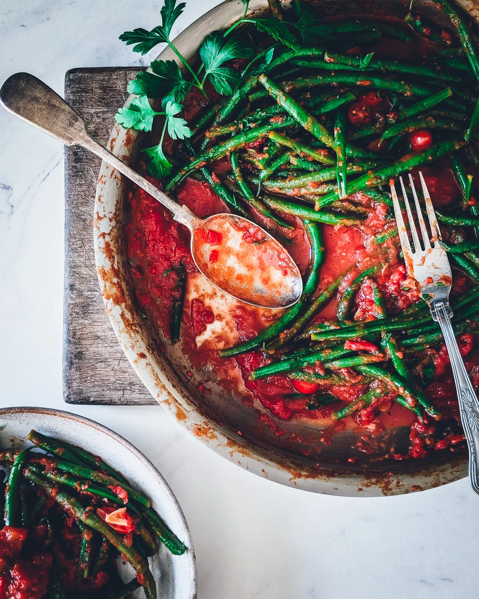 Pan with green beans in garlicky tomato sauce on a wooden chopping board. The pan is half empty, with some cutlery in it. Next to the pan a small dish full of green beans.