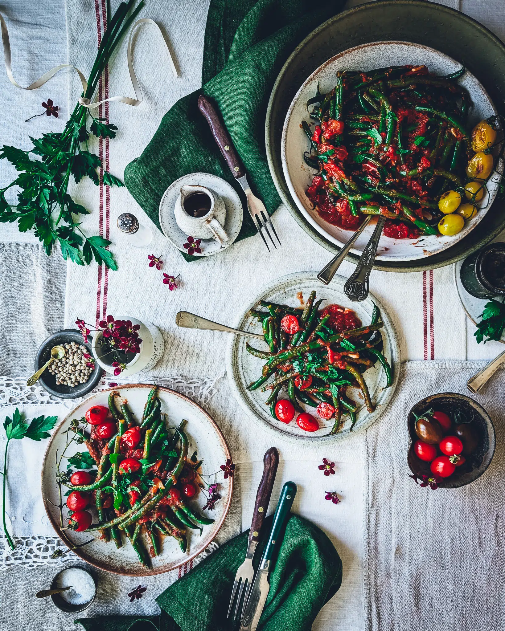Table with three plates full of green beans in garlicky tomato sauce. On the table also some green napkins, cutlery, cherry tomatoes, small purple flowers and various trinkets.