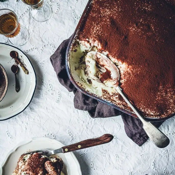 Tin of tiramisu partially eaten. A large serving spoon is in the tin. On the side a plate with tiramisu on it.