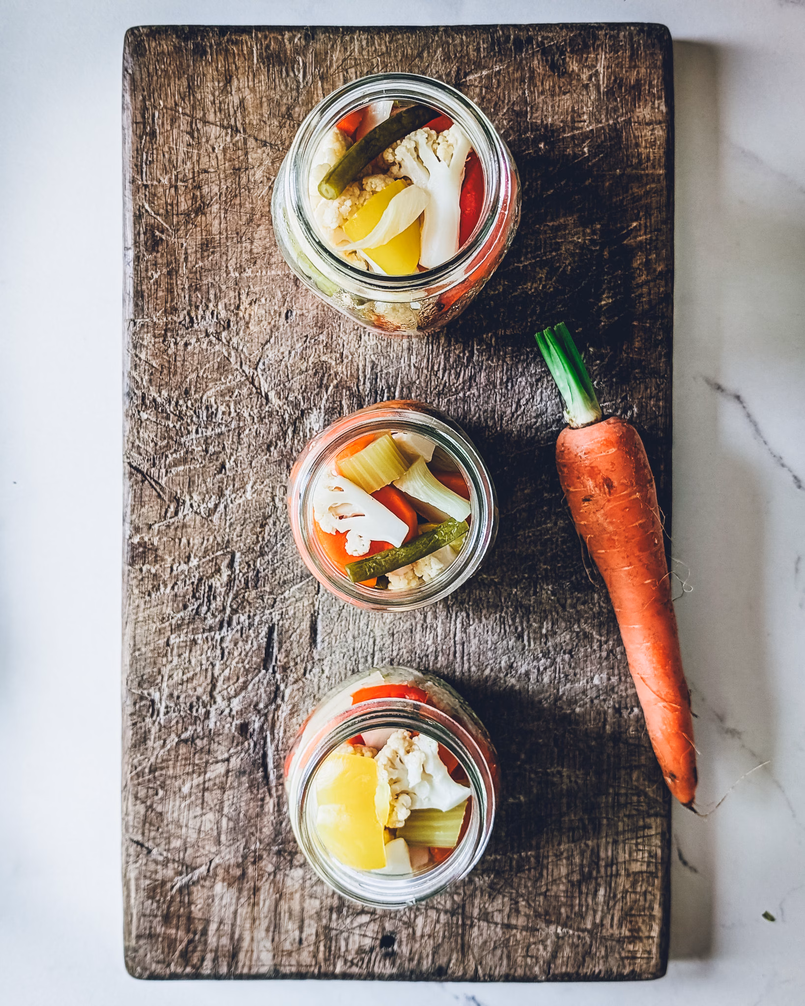 Three jars of pickled vegetables on a wooden chopping board. A carrot is also on the chopping board.