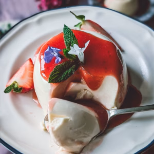 A beautifully styled authentic Italian panna cotta with strawberry sauce turned out on a vintage ceramic plate, topped with vibrant violas.