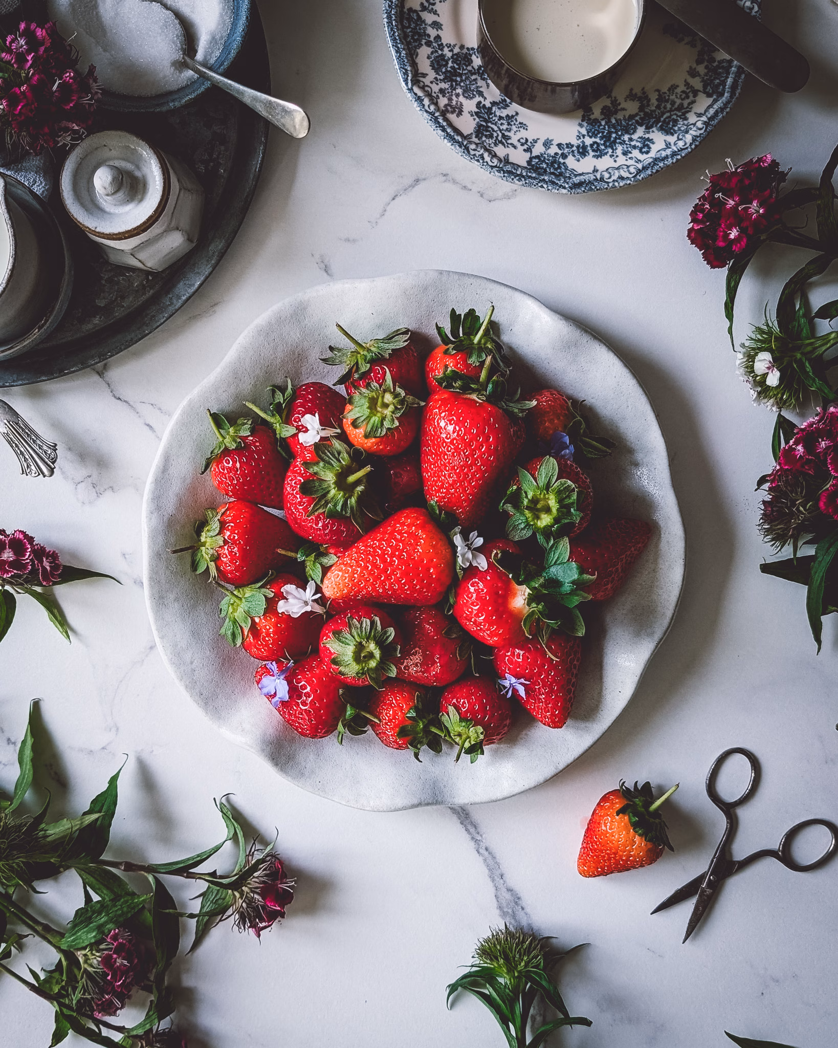 Plate full of fresh strawberries. On the table there are also several trinkets full of cream, sugar, milk, as well as some purple flowers.