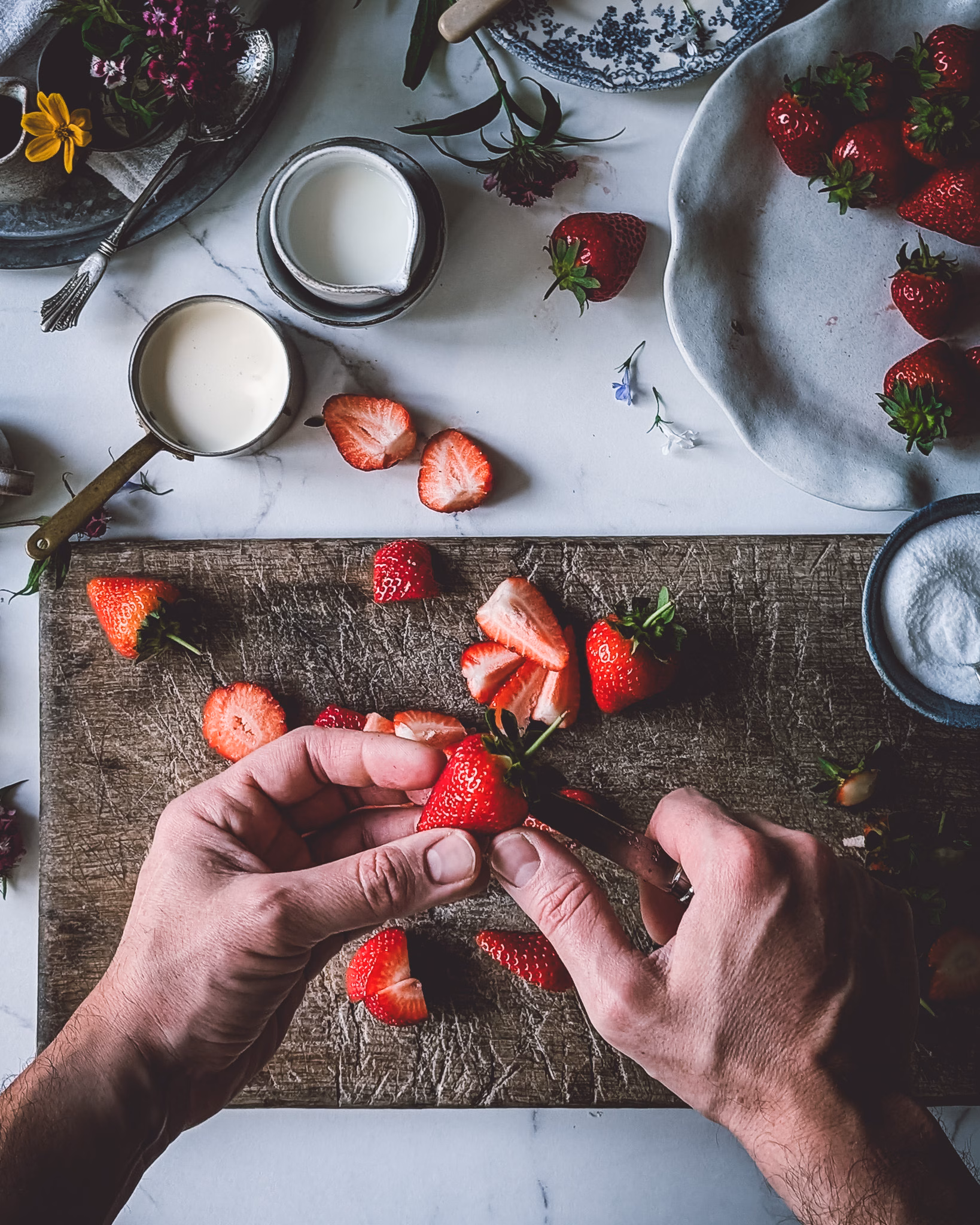Chopping board with chopped strawberries and a knife, next to a plate full of whole strawberries and a small pan with cream. A person is chopping a strawberry.