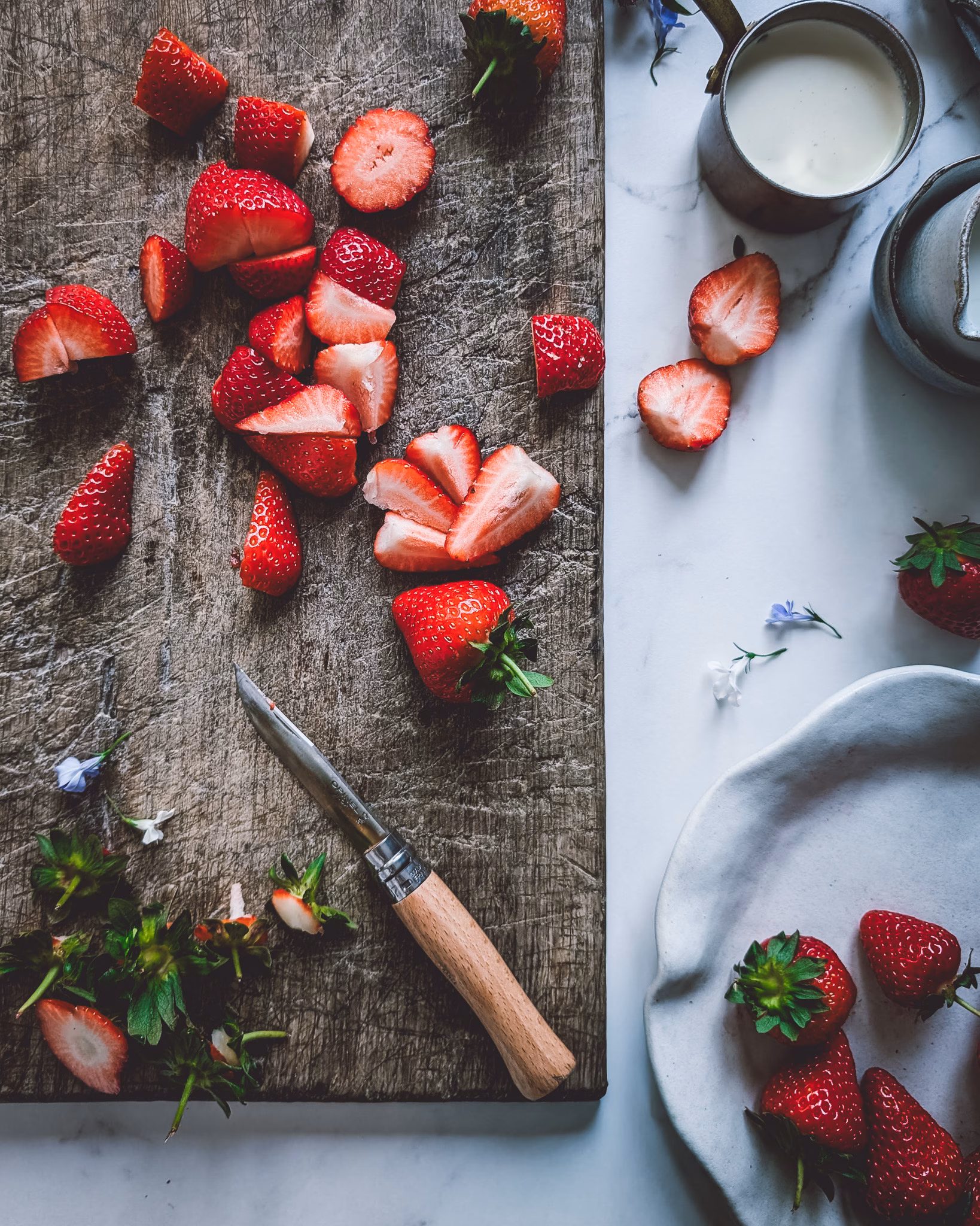 Chopping board with chopped strawberries and a knife, next to a plate full of whole strawberries and a small pan with cream