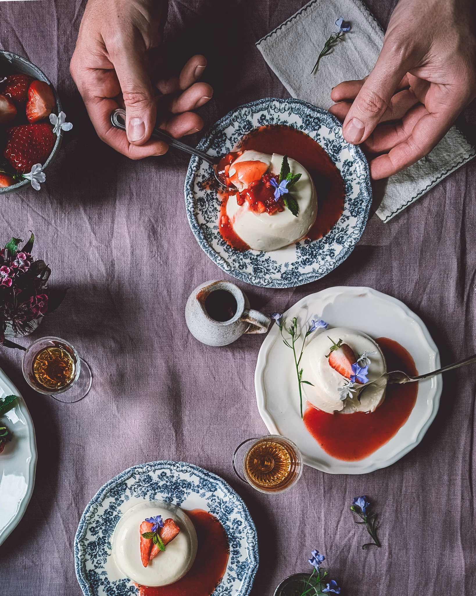 Plates with panna cotta with strawberry sauce decorated with flowers. On the table there are also trinkets and glasses, alongside cutlery and napkins. A person is eating from one of the plates.