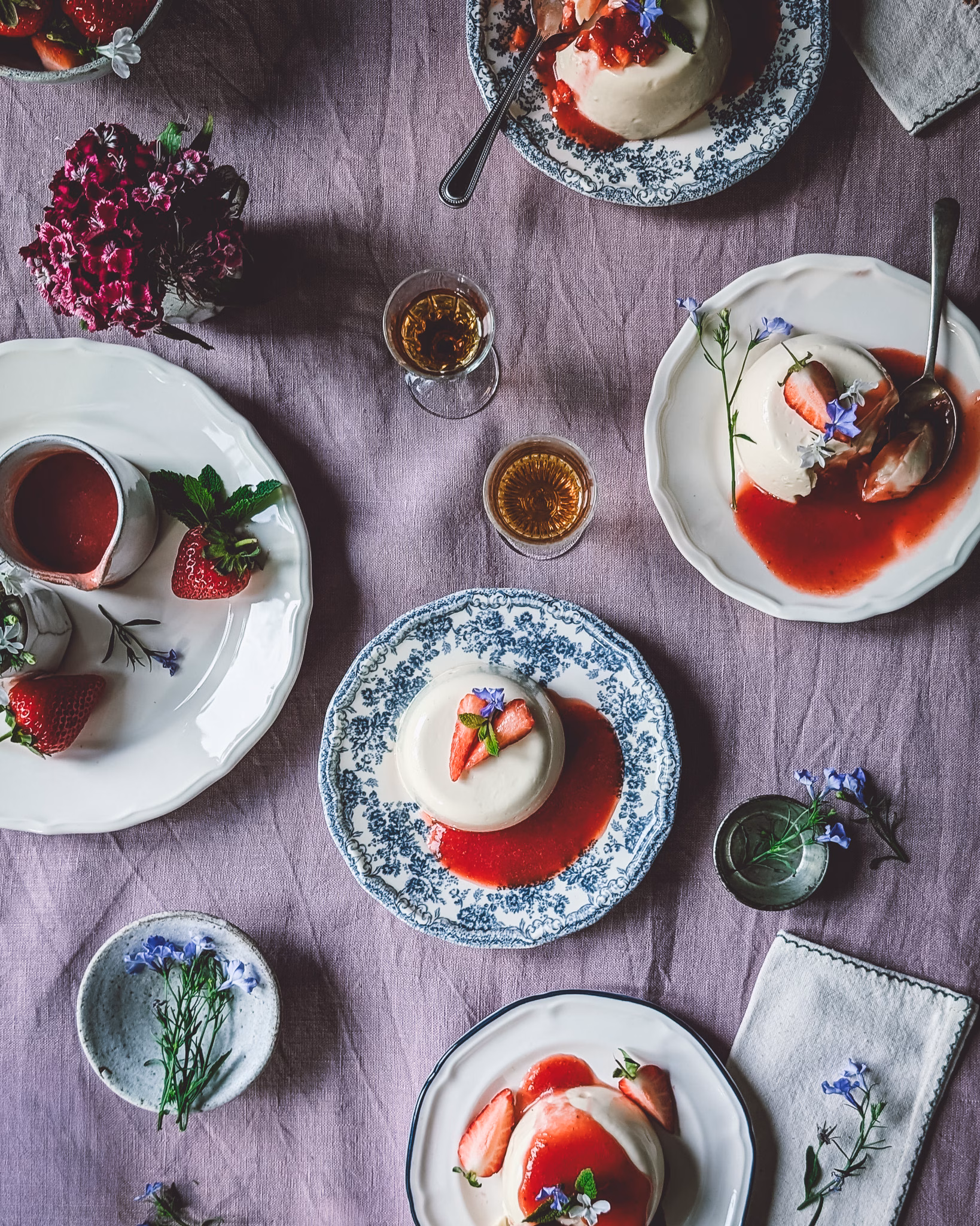 Plates with panna cotta with strawberry sauce decorated with flowers. On the table there are also trinkets and glasses, alongside cutlery and napkins.