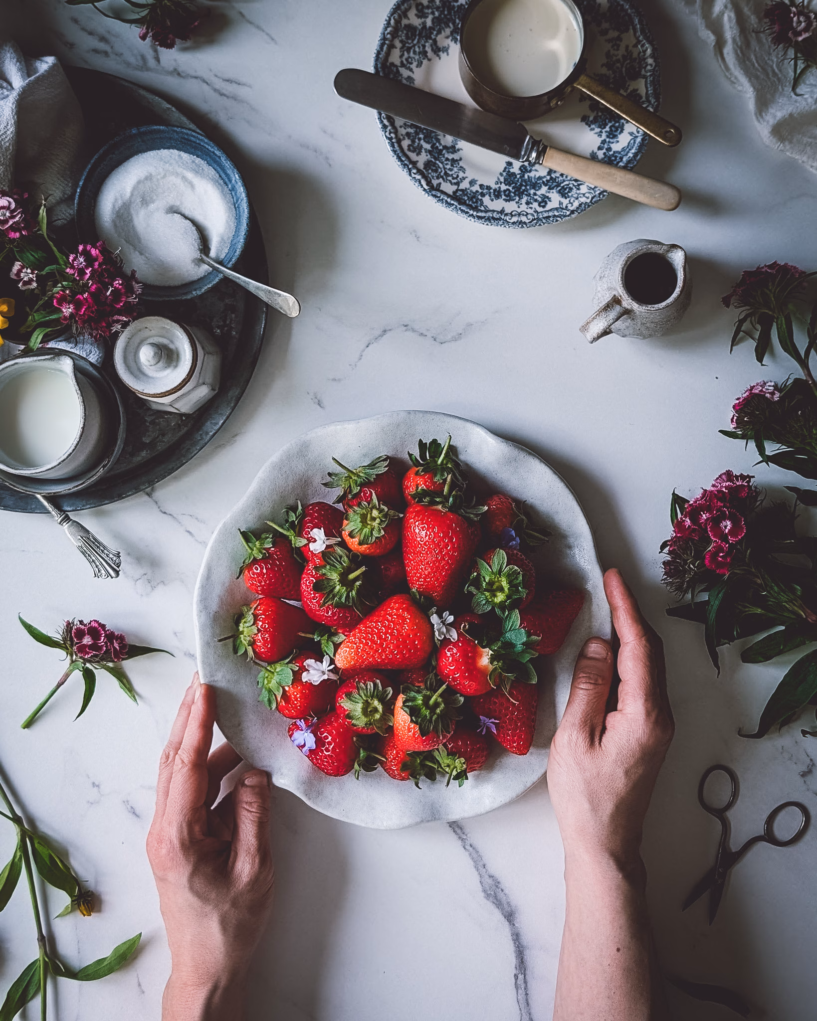 Hands of person holding onto plate full of fresh strawberries. On the table there are also several trinkets full of cream, sugar, milk, as well as some purple flowers.