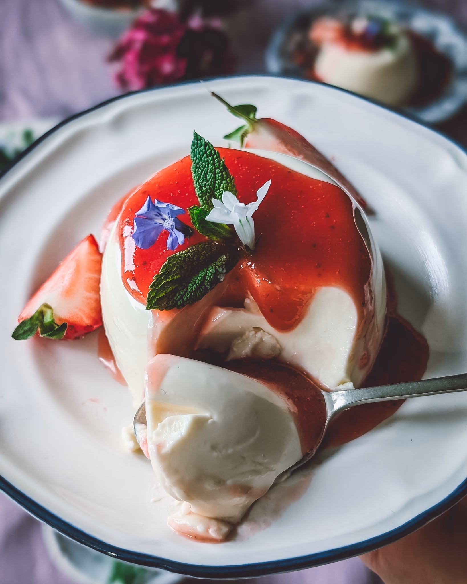 A beautifully styled authentic Italian panna cotta with strawberry sauce turned out on a vintage ceramic plate, topped with vibrant violas.