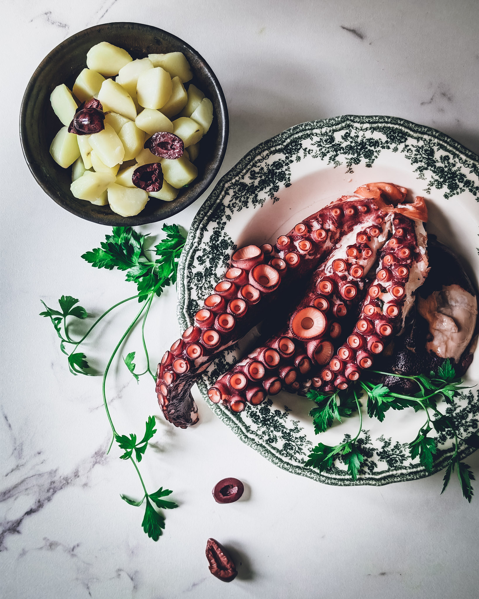 Cooked octopus on a plate, next to a plate with cook potatoes and some olives and parsley