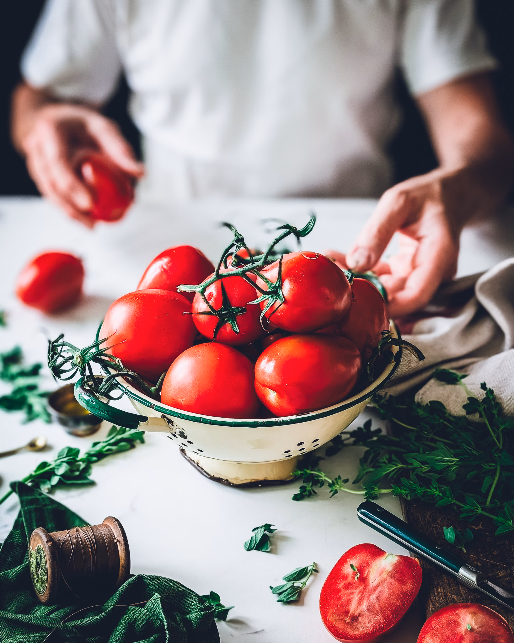 A vintage enamel colander filled with fresh, bright red vine tomatoes on a kitchen counter, with a person in a white shirt reaching for them in the blurred background.