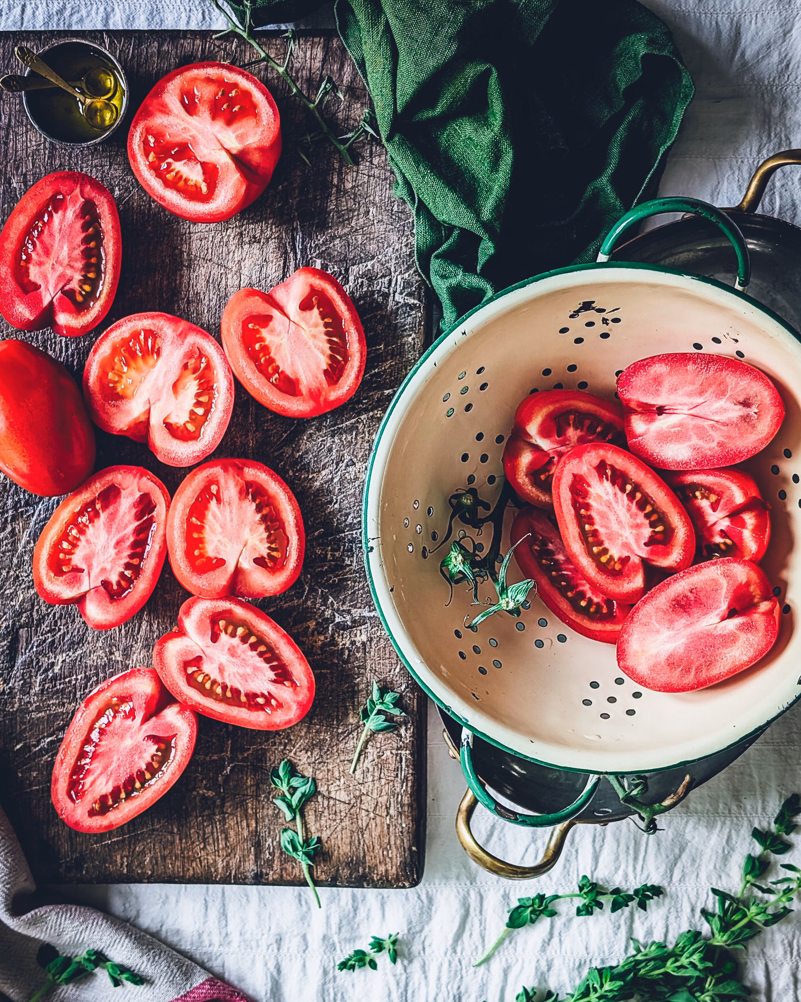 Table with a colander and a chopping board covered in tomatoes chopped in half. On the table there is also a green napkin and scattered oregano.