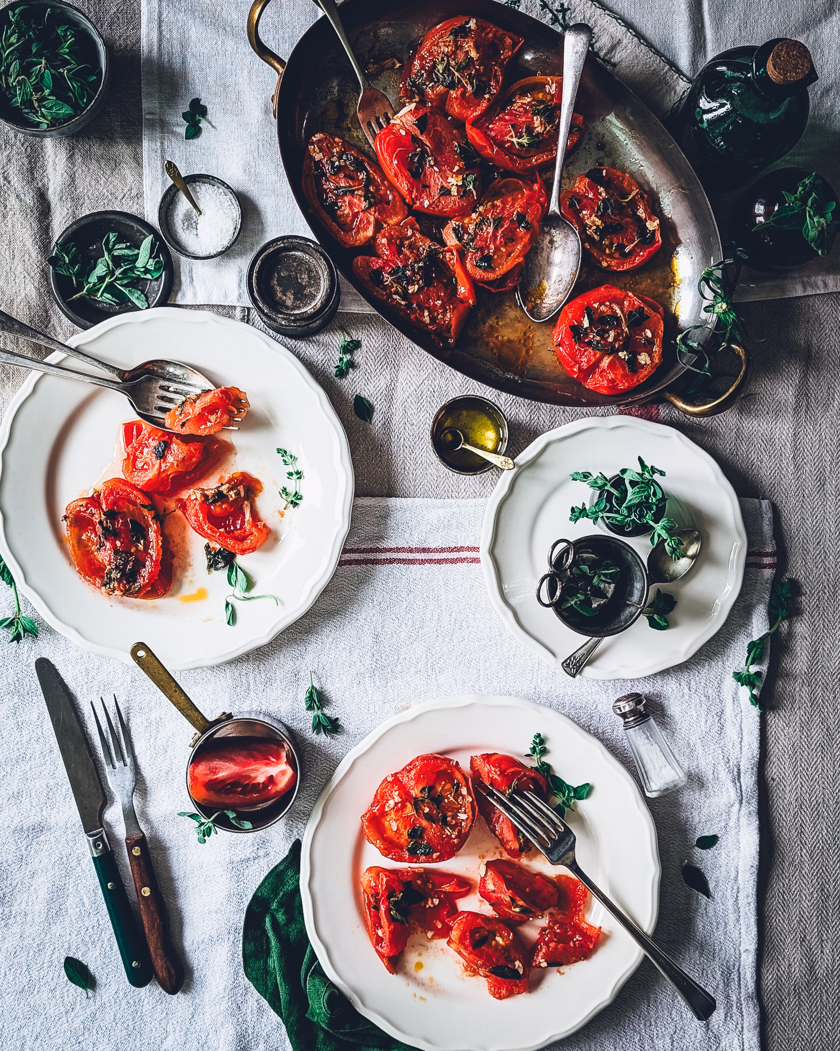 An overhead view of a beautifully set rustic table featuring pan-fried red tomatoes with garlic and herbs. A large oval copper platter sits at the top filled with cooked tomatoes, while two white ceramic plates below display portions of the tomatoes. The scene is accented by small bowls of fresh herbs, oil, a salt shaker, and vintage silverware on a textured linen tablecloth.