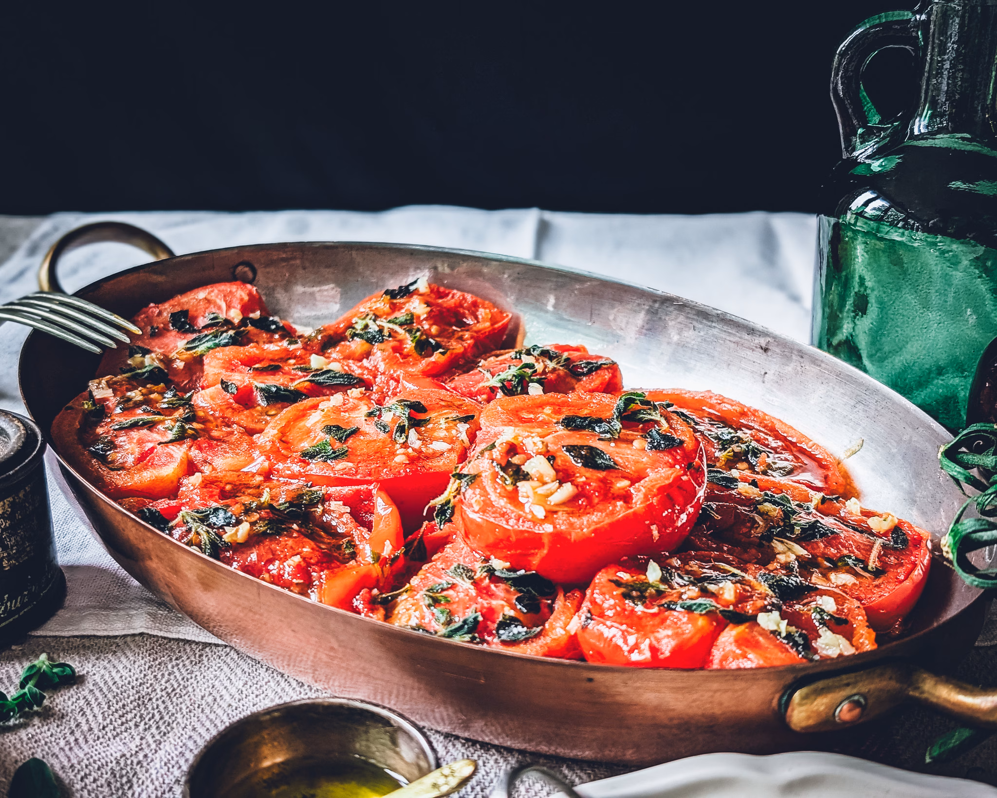 Pan of pan-fried tomatoes with fresh garden herbs on a table covered in a white tablecloth. Next to the pan some trinkets with salt and oil, fresh oregano and two empty places and cutlery.