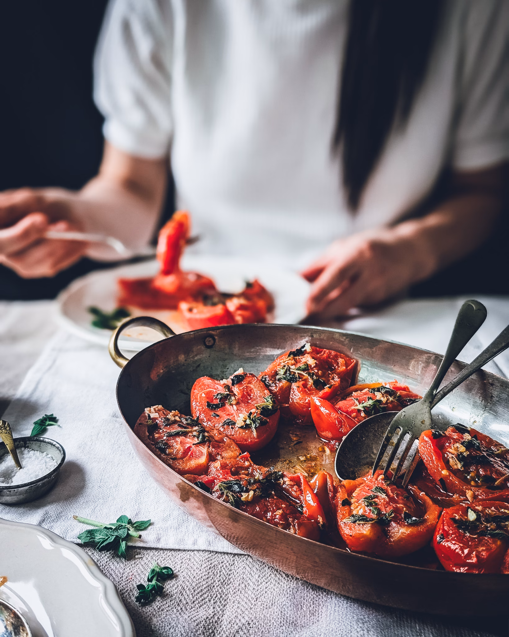 Pan of pan-fried tomatoes on a table covered in a white tablecloth. Behind the pan a woman is eating tomatoes from a white scalloped plate.