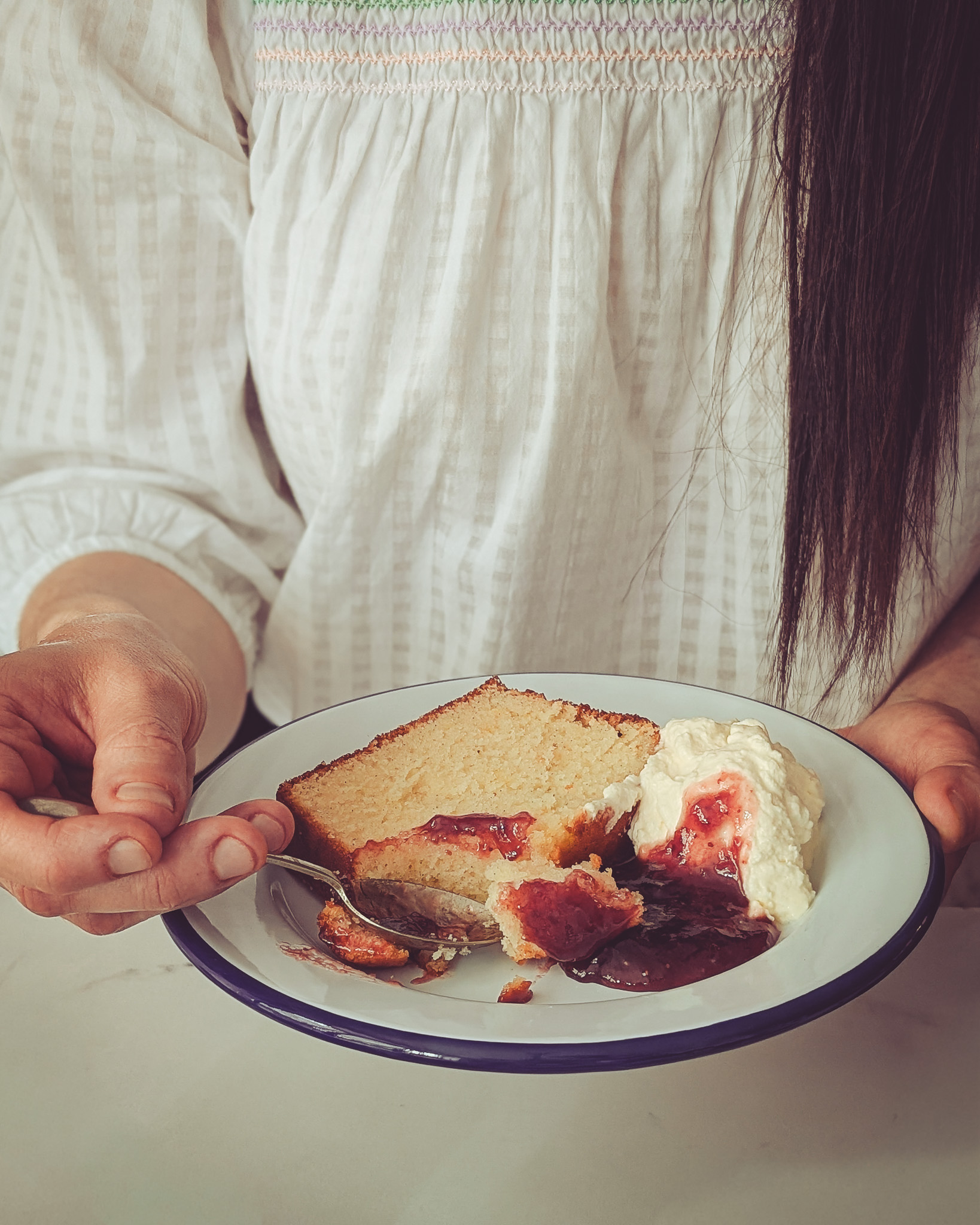 Woman eating a slice of yoghurt and rose loaf cake served with whipped cream and jam.