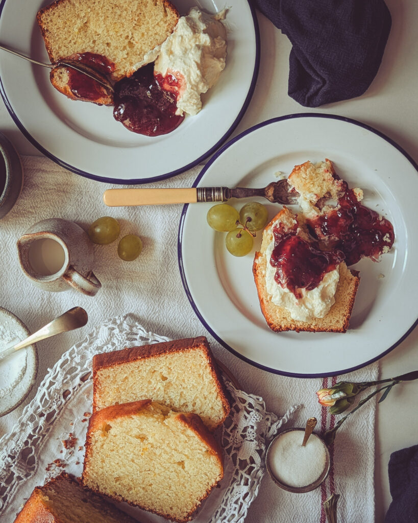Two plates with slices of yoghurt and rose loaf cake (Italian plumcake) served alongside whipped cream and jam.