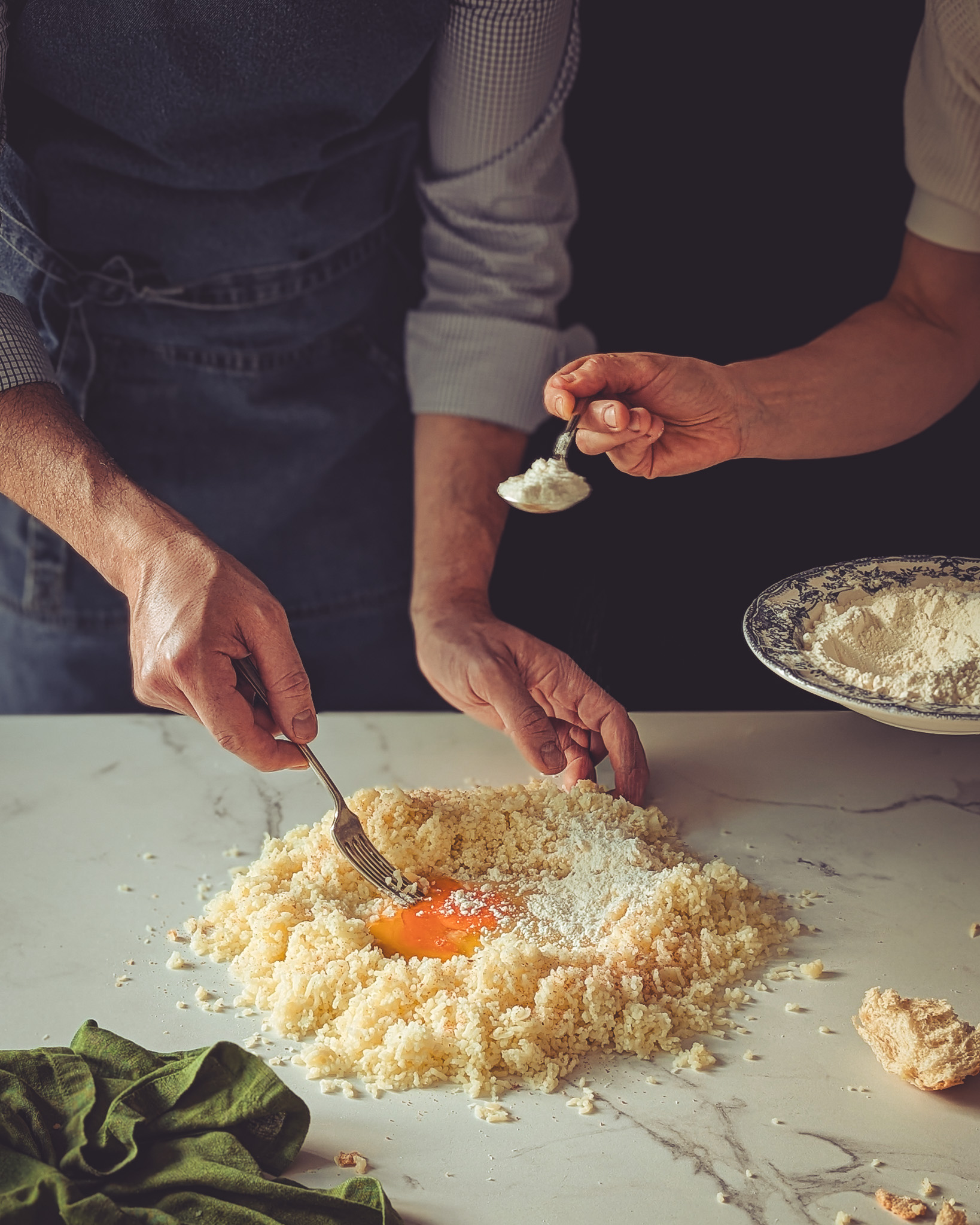 Woman pouring flour on a table while a man is mixing it with eggs.