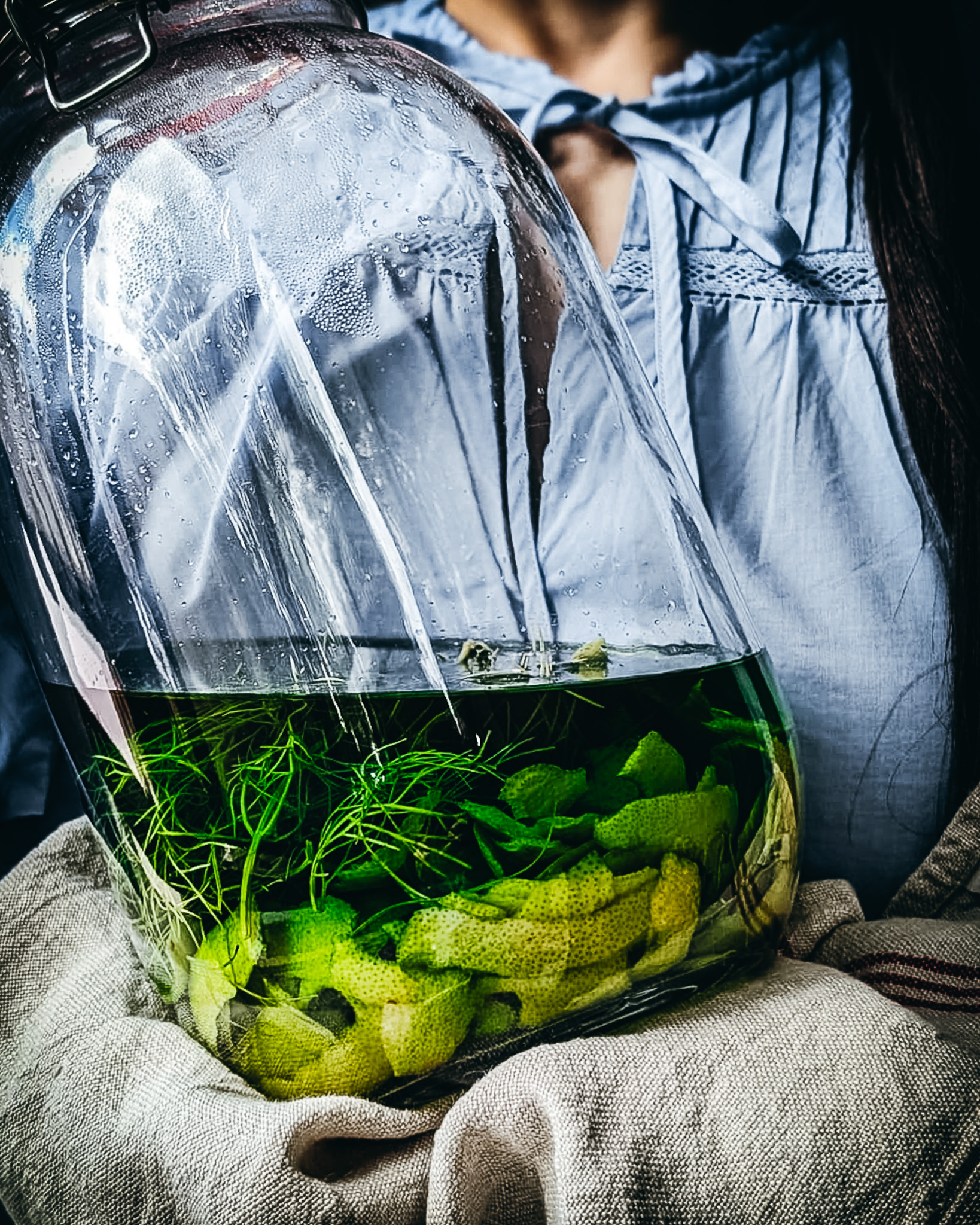 Woman holding a large jar full of lemon peels and fennel fronds infusing in alcohol for homemade limoncello.
