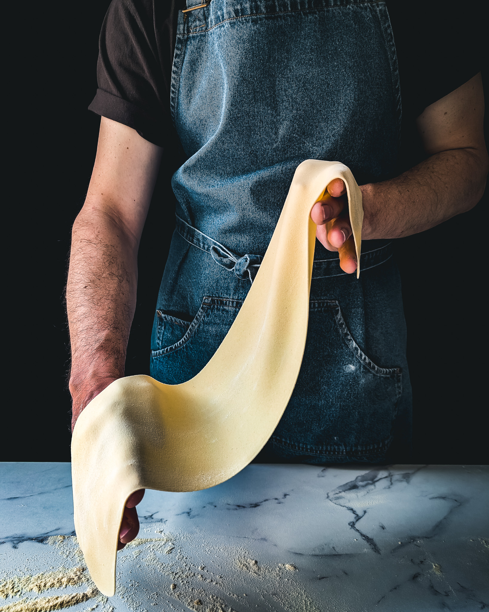 Person wearing blue apron and holding sheet of fresh pasta.